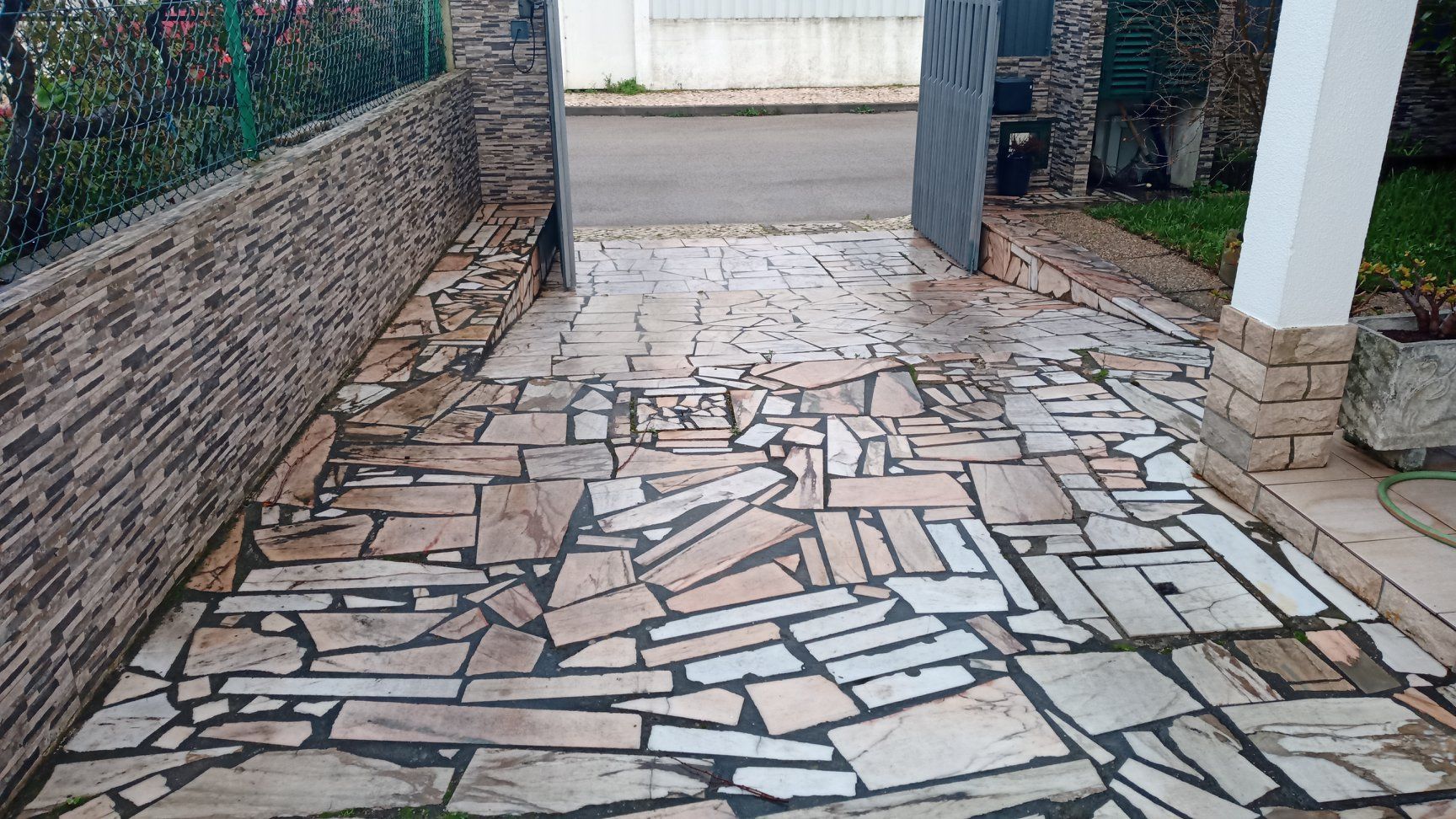 Entrada de automóveis pavimentada com pedra clara, ladeada por um muro de pedra empilhada e uma coluna branca, que dá acesso a uma rua.