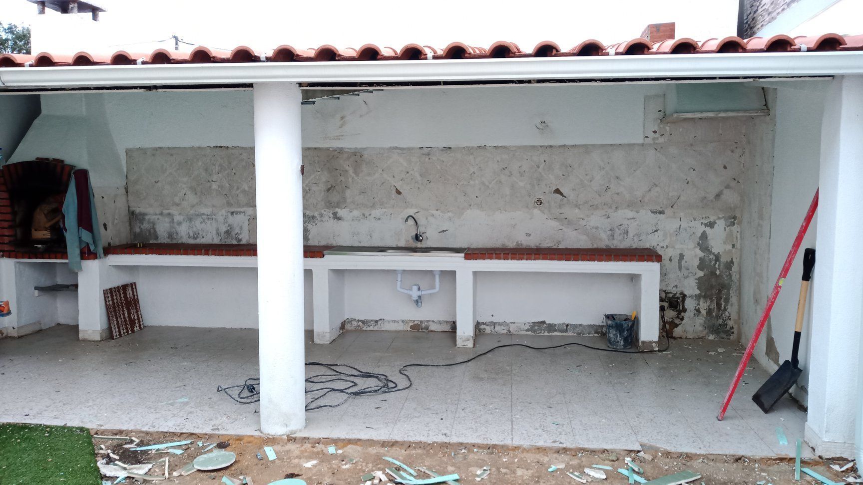 Cozinha externa com paredes e bancadas brancas, churrasqueira e danos aparentes na parede.