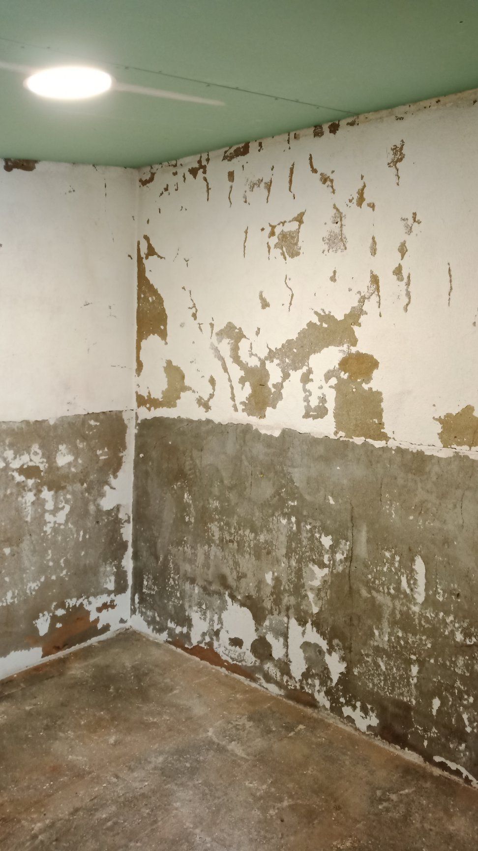 Tinta descascando e descoloração nos cantos internos das paredes; iluminação fraca, piso de concreto, teto verde.