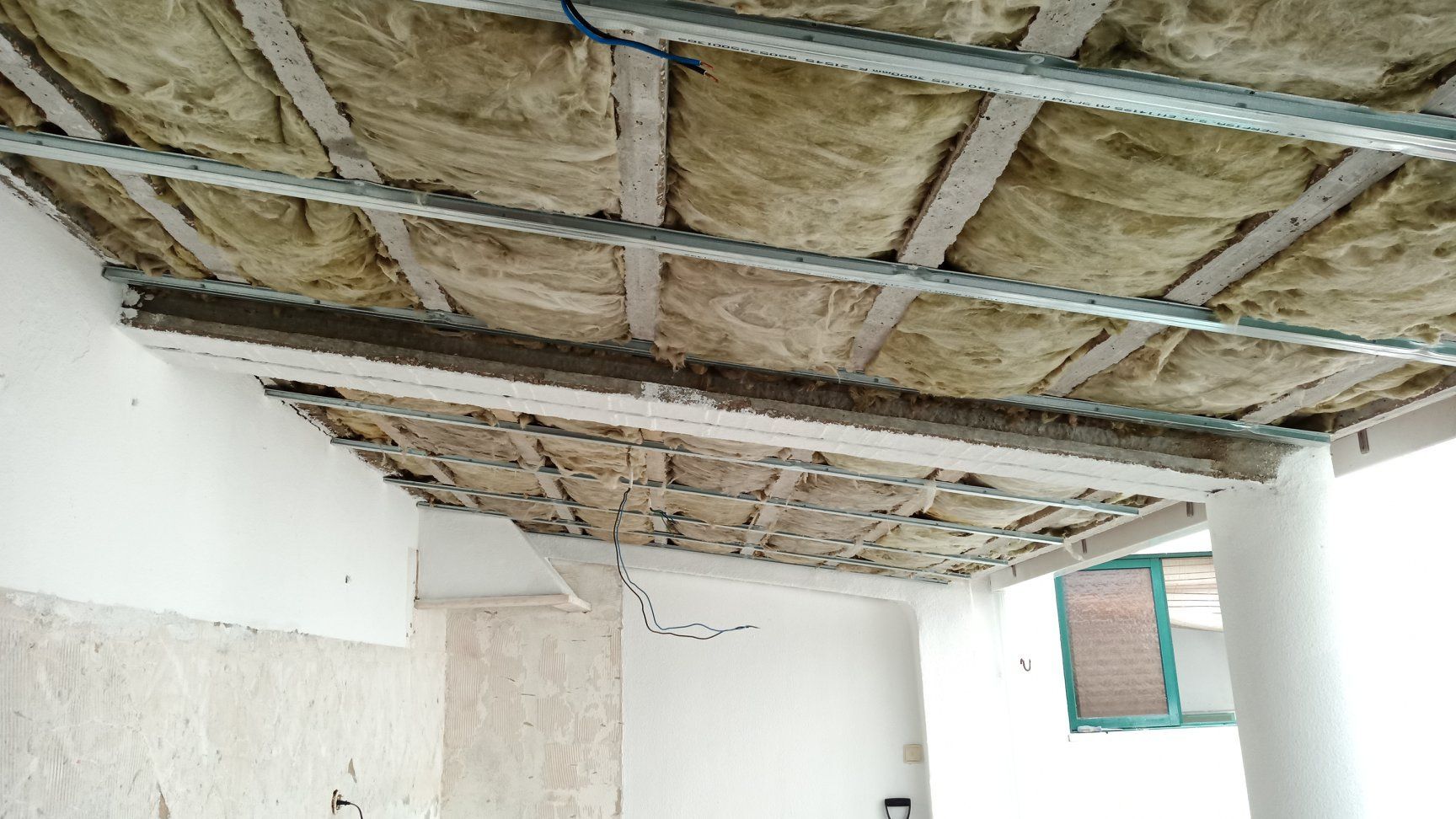 Construção do teto em andamento, mostrando o isolamento exposto entre as estruturas metálicas, dentro de uma sala.