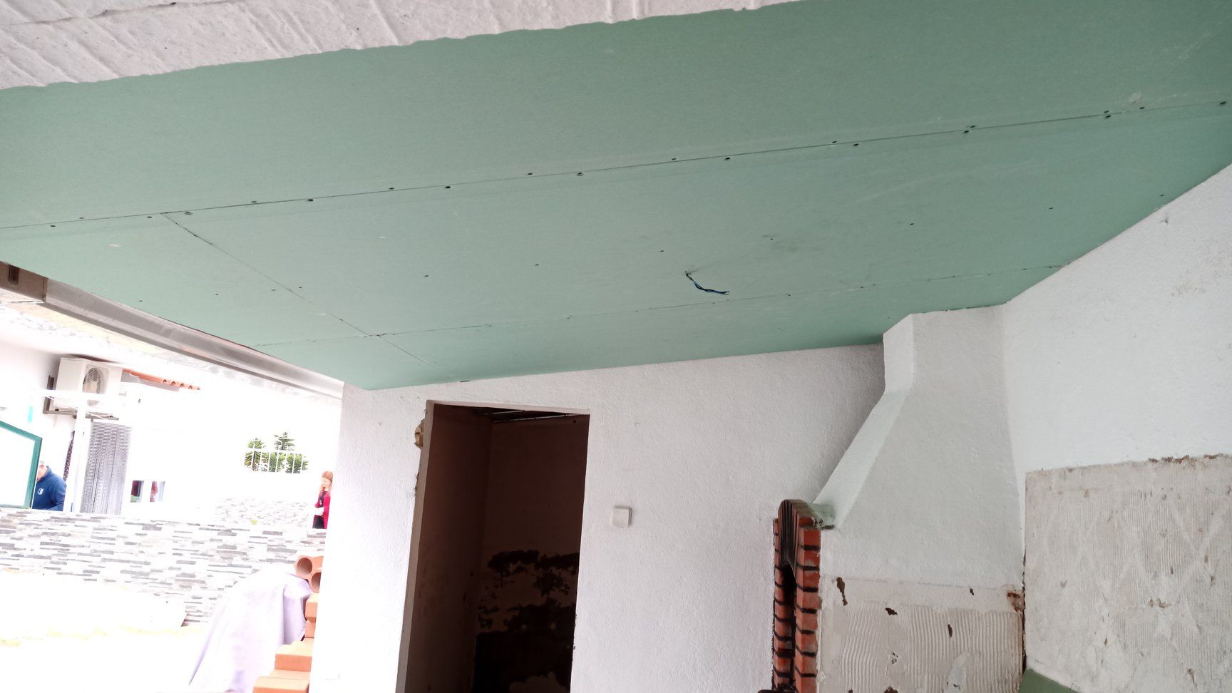 Teto pintado de verde com juntas visíveis; cômodo inacabado com paredes brancas, porta e vista para o exterior.