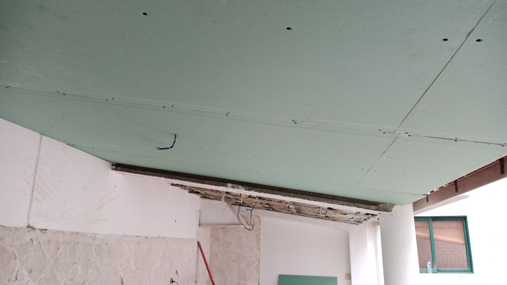 Instalação de forro de gesso acartonado verde em um cômodo. Vigas expostas e paredes parcialmente acabadas visíveis.