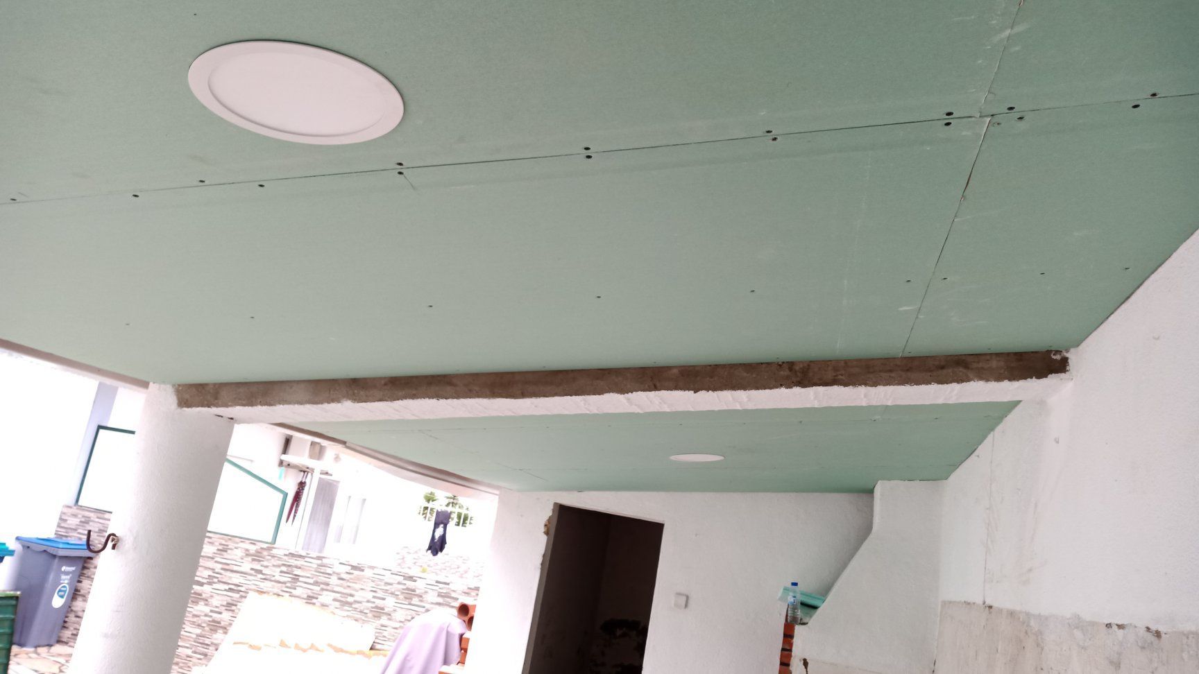 Teto verde com luminária circular, viga de concreto e paredes brancas. Ambiente externo.