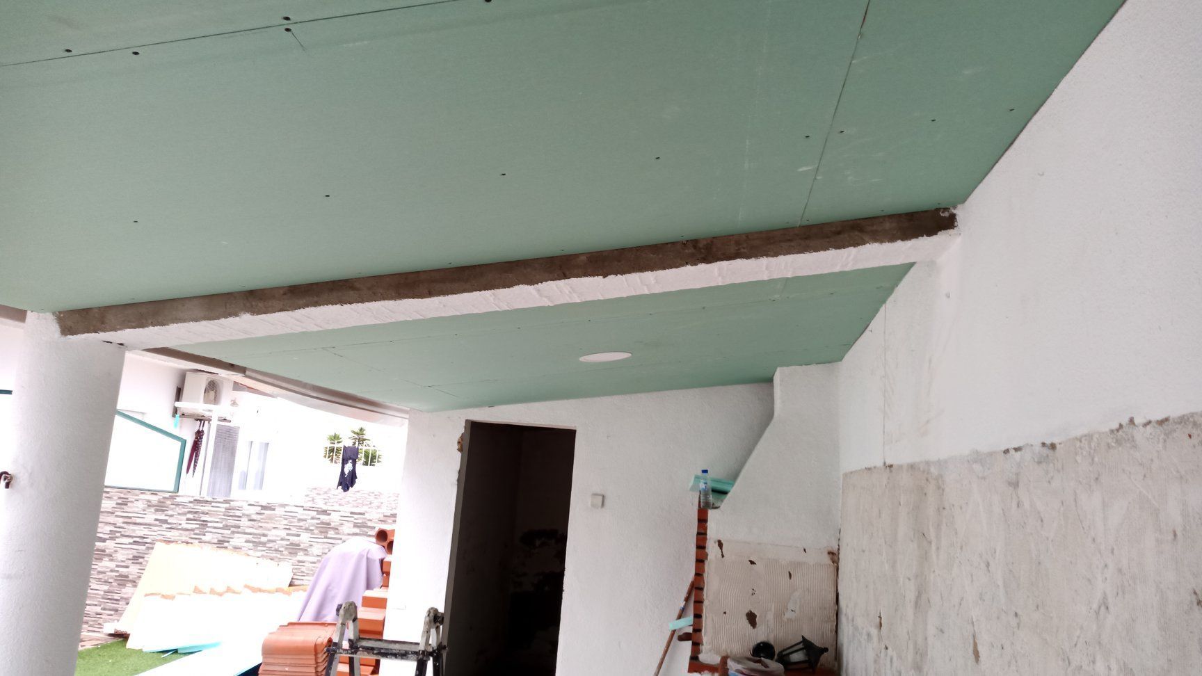 Teto de gesso cartonado verde com vigas de concreto aparentes, paredes brancas, porta, ambiente externo.