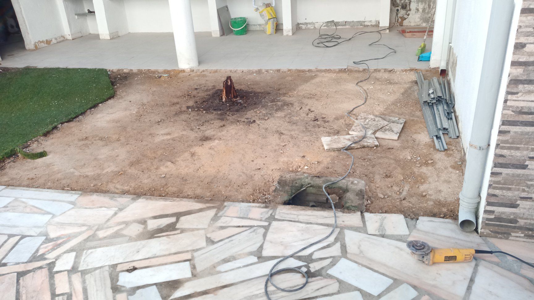 Um canteiro de obras com terra exposta e um pátio de pedra. Um toco de árvore está no centro.