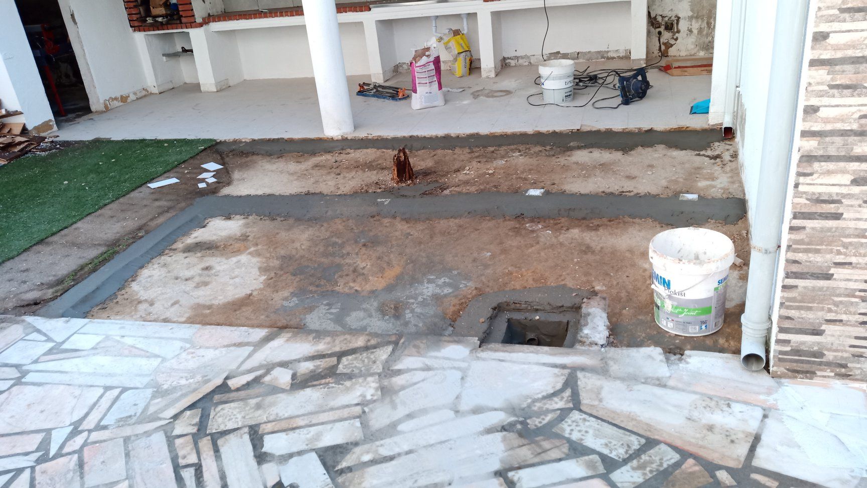 Canteiro de obras: pátio inacabado com estrutura de concreto, balde e sistema de drenagem.