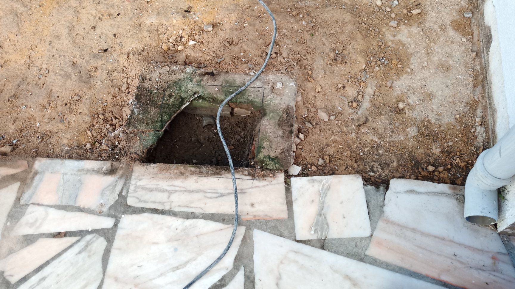 Buraco na terra, perto de um pátio de pedra e de uma parede branca com calha. Um fio entra no buraco.