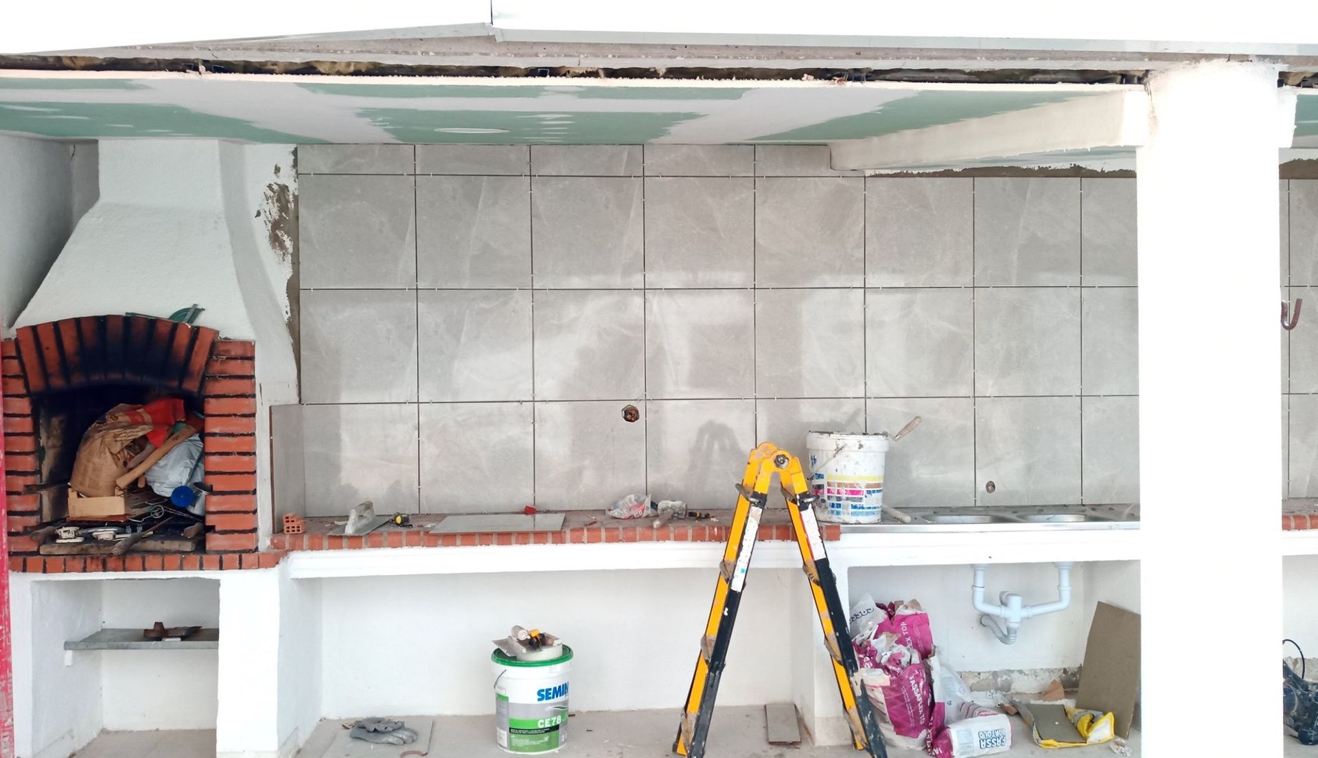 Cozinha externa em construção, com backsplash de azulejos, lareira, escada e suprimentos.