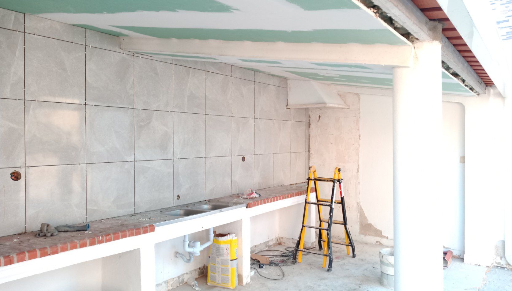 Construção de uma cozinha externa; paredes revestidas com azulejos brancos, bancada de tijolos, escada, teto inacabado.