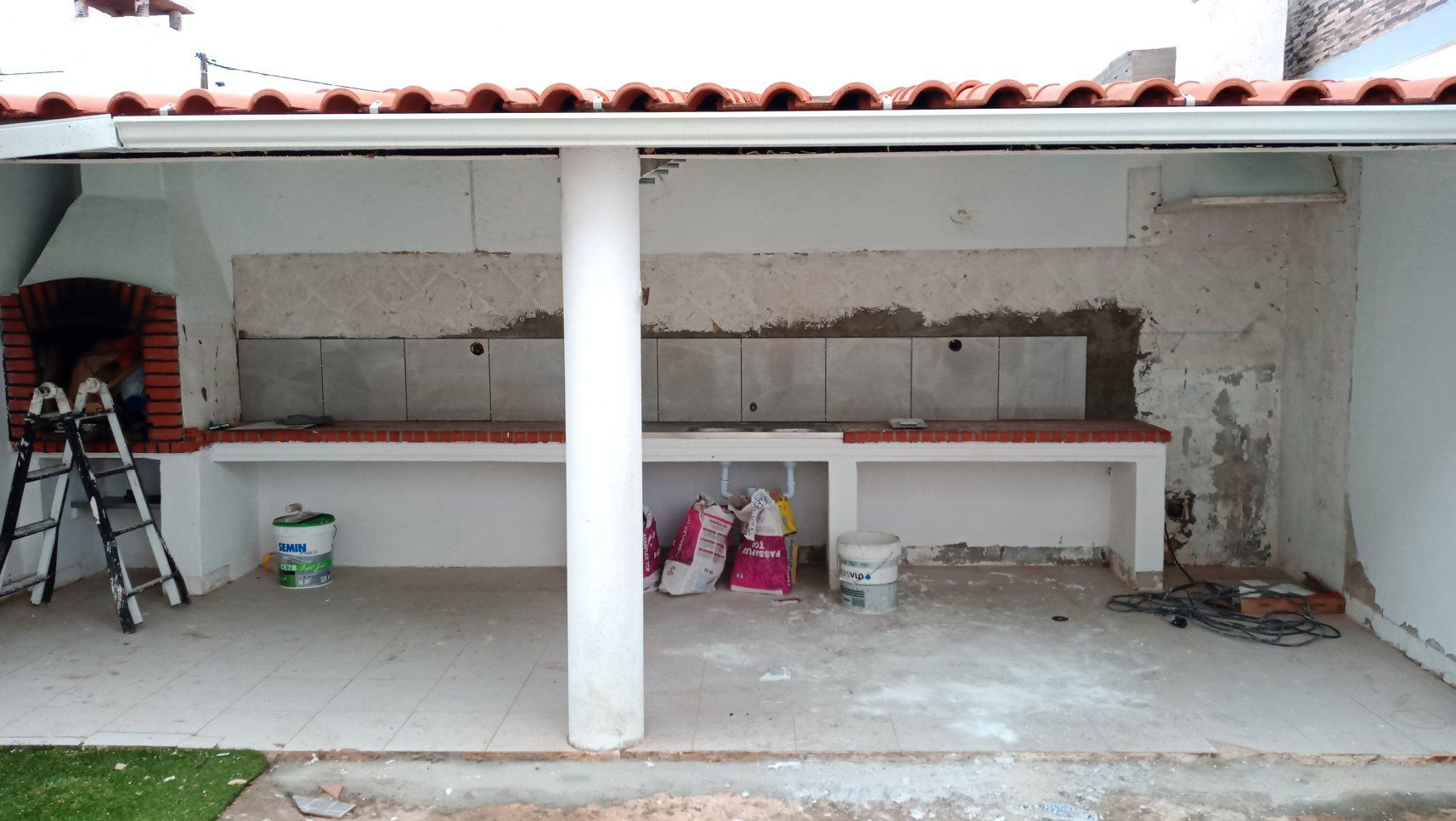 Cozinha externa em construção, com churrasqueira de tijolos, bancadas de concreto e paredes inacabadas.