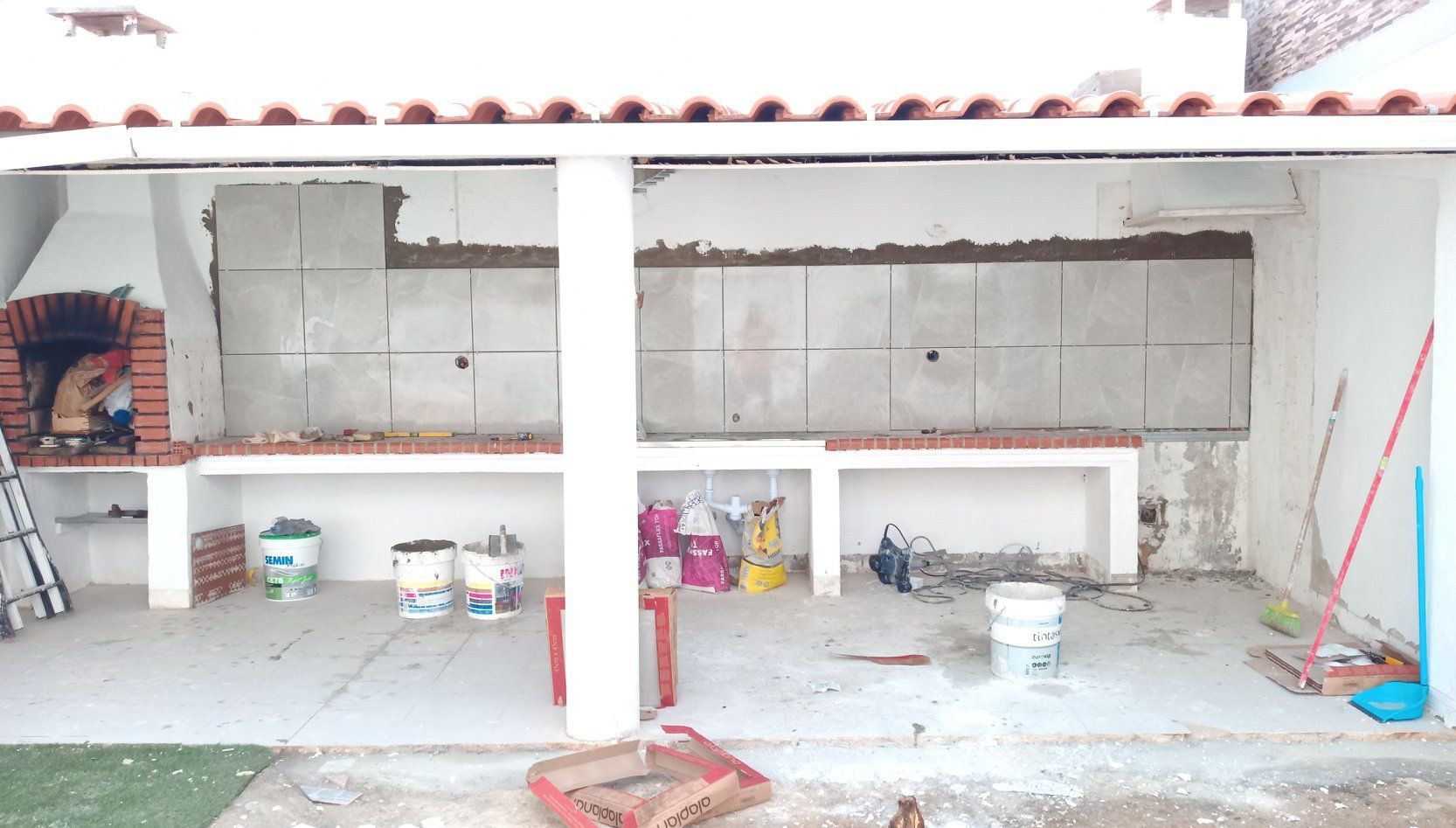 Cozinha externa em construção; churrasqueira de tijolos, bancadas de concreto, paredes cinza, ferramentas e suprimentos.