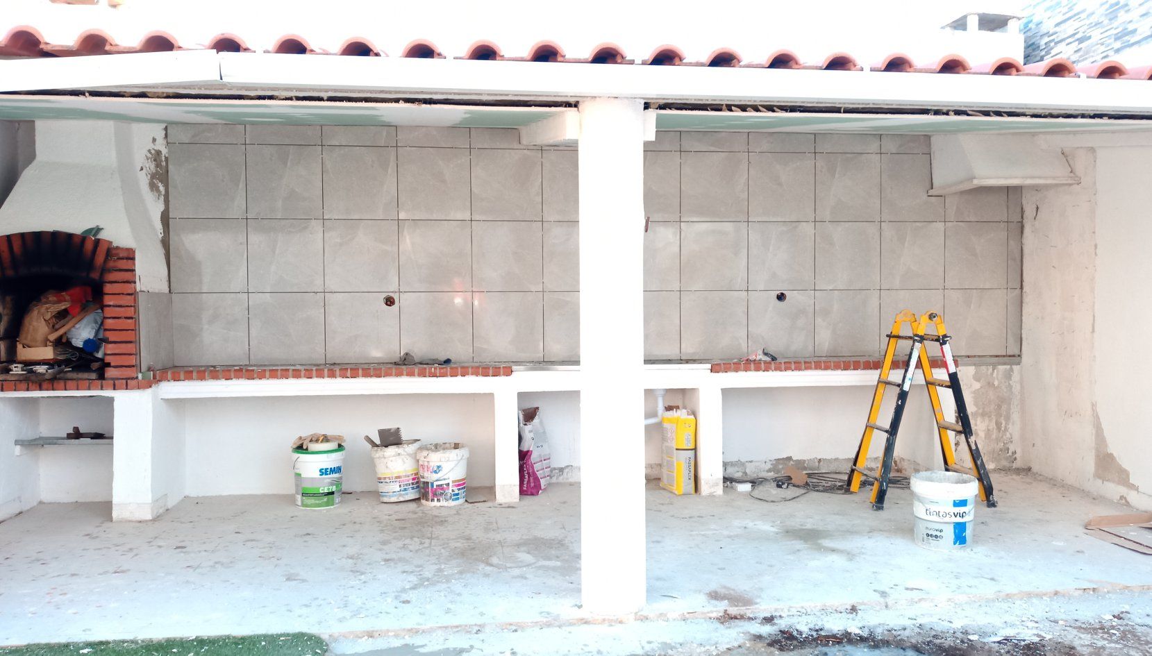 Cozinha externa em construção: churrasqueira de tijolos, bancadas de concreto, paredes inacabadas, ferramentas, latas de tinta.