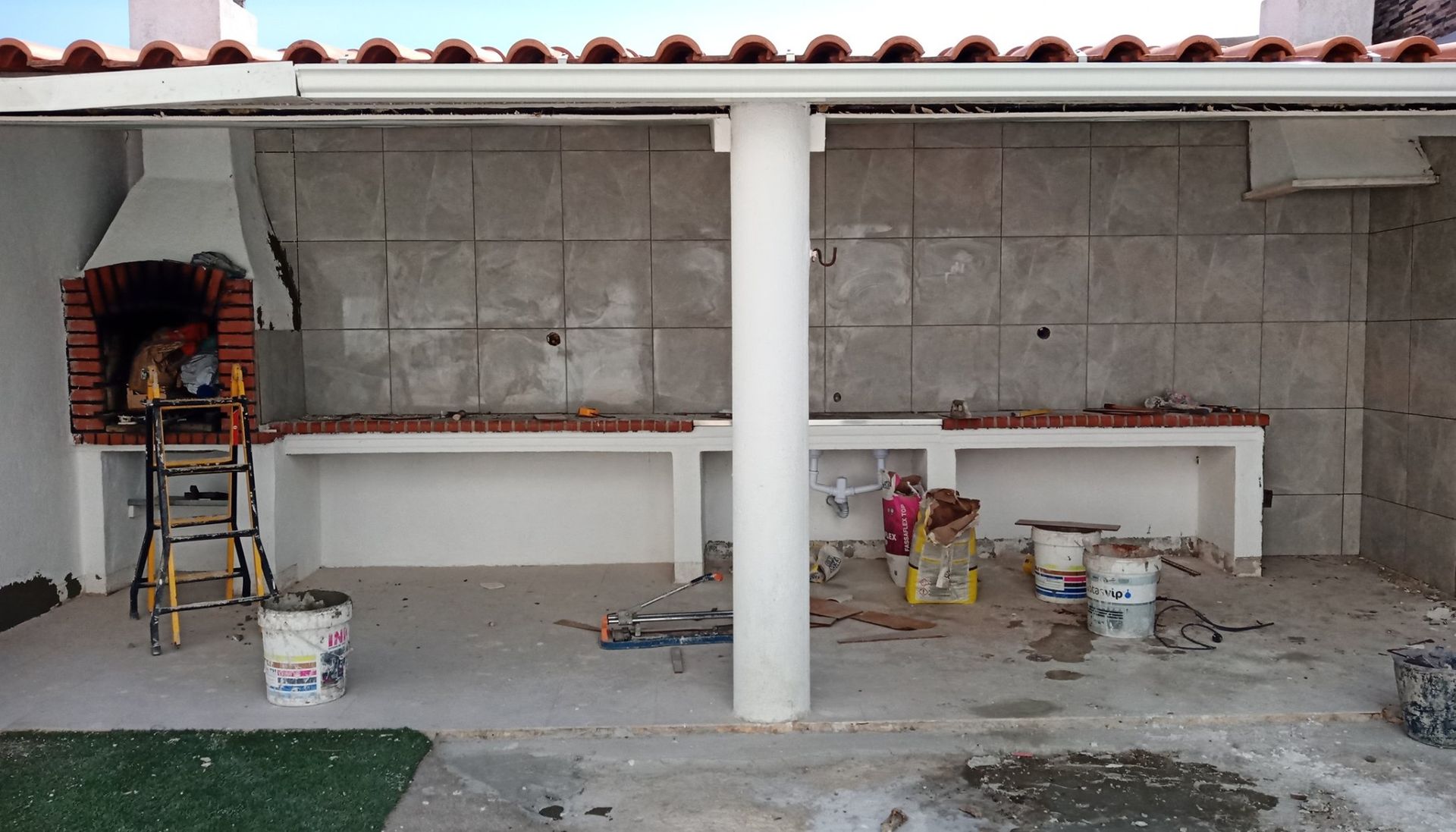 Cozinha externa em construção, com churrasqueira de tijolos, parede revestida com azulejos e utensílios espalhados pelo chão de concreto.