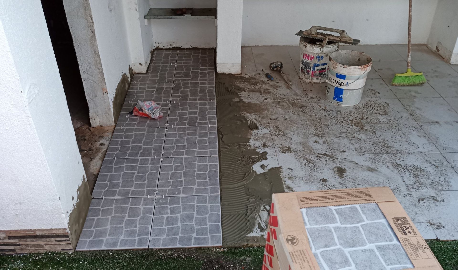 Instalação de ladrilhos em um pátio. É possível ver ladrilhos de pedra cinza, argamassa, ferramentas e um piso parcialmente revestido.