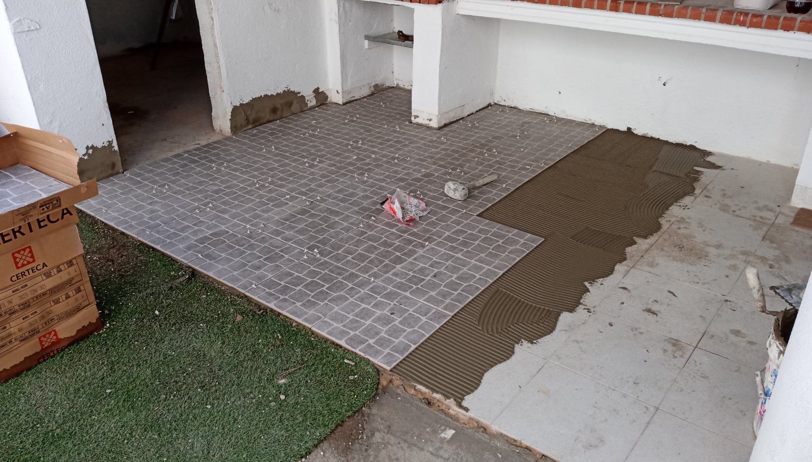 Assentamento de azulejos no piso de um pátio; azulejos cinza, adesivo e caixa de azulejos visíveis.