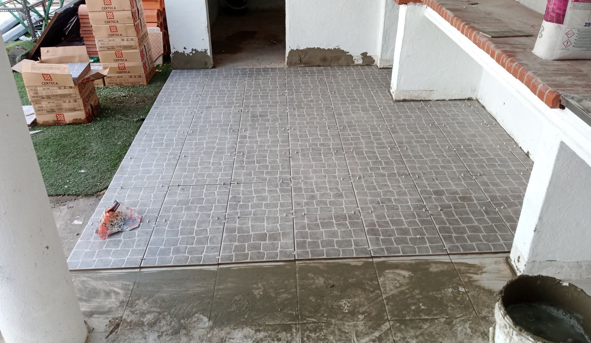 Instalação de novo piso de ladrilho cinza em um pátio, com materiais e construção em andamento.