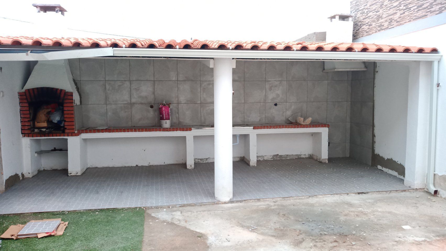 Área externa para churrasco com piso de concreto, lareira de tijolos e paredes revestidas com azulejos.