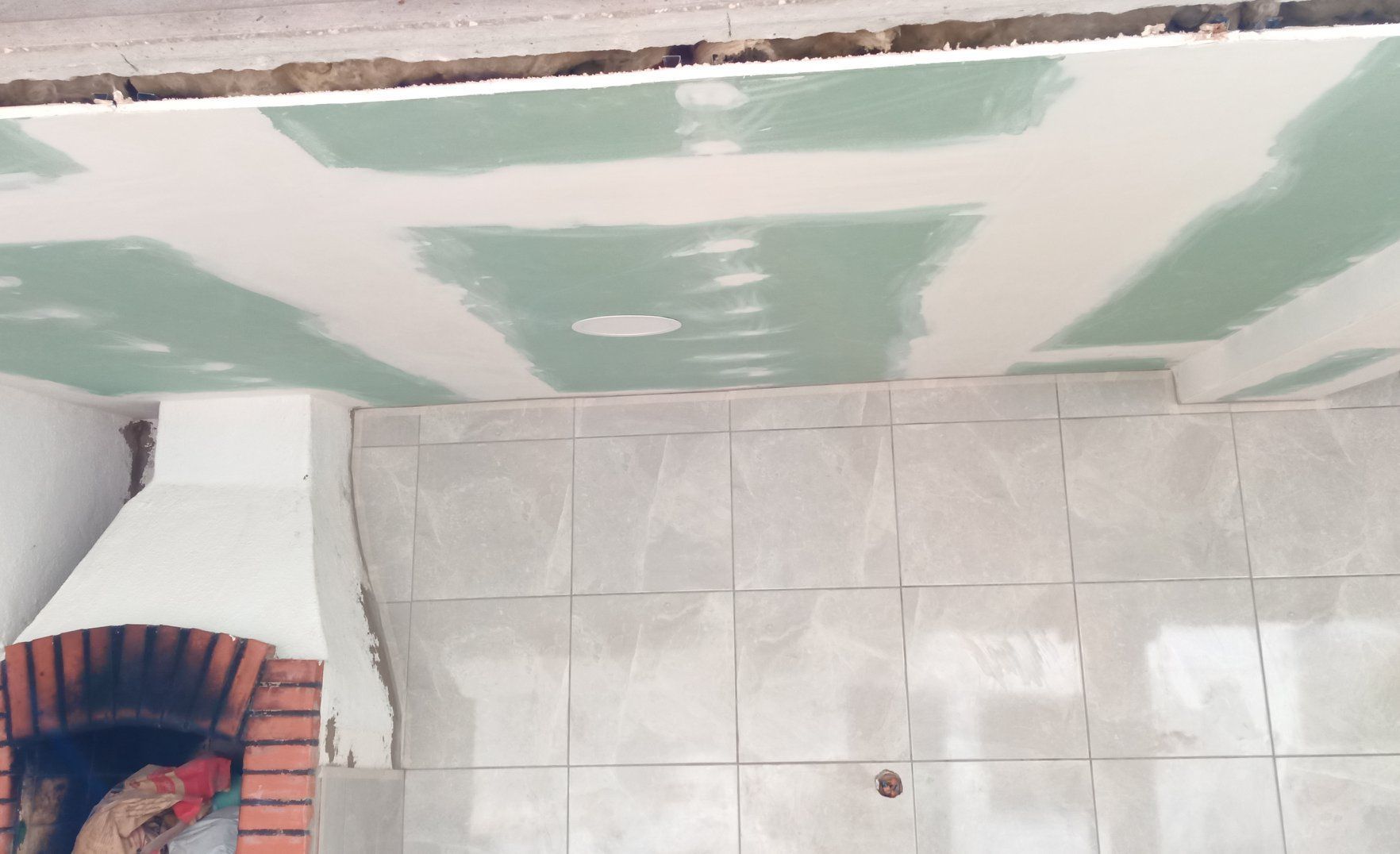 Instalação de drywall no teto em andamento acima da parede revestida com azulejos, próximo a uma lareira de tijolos.