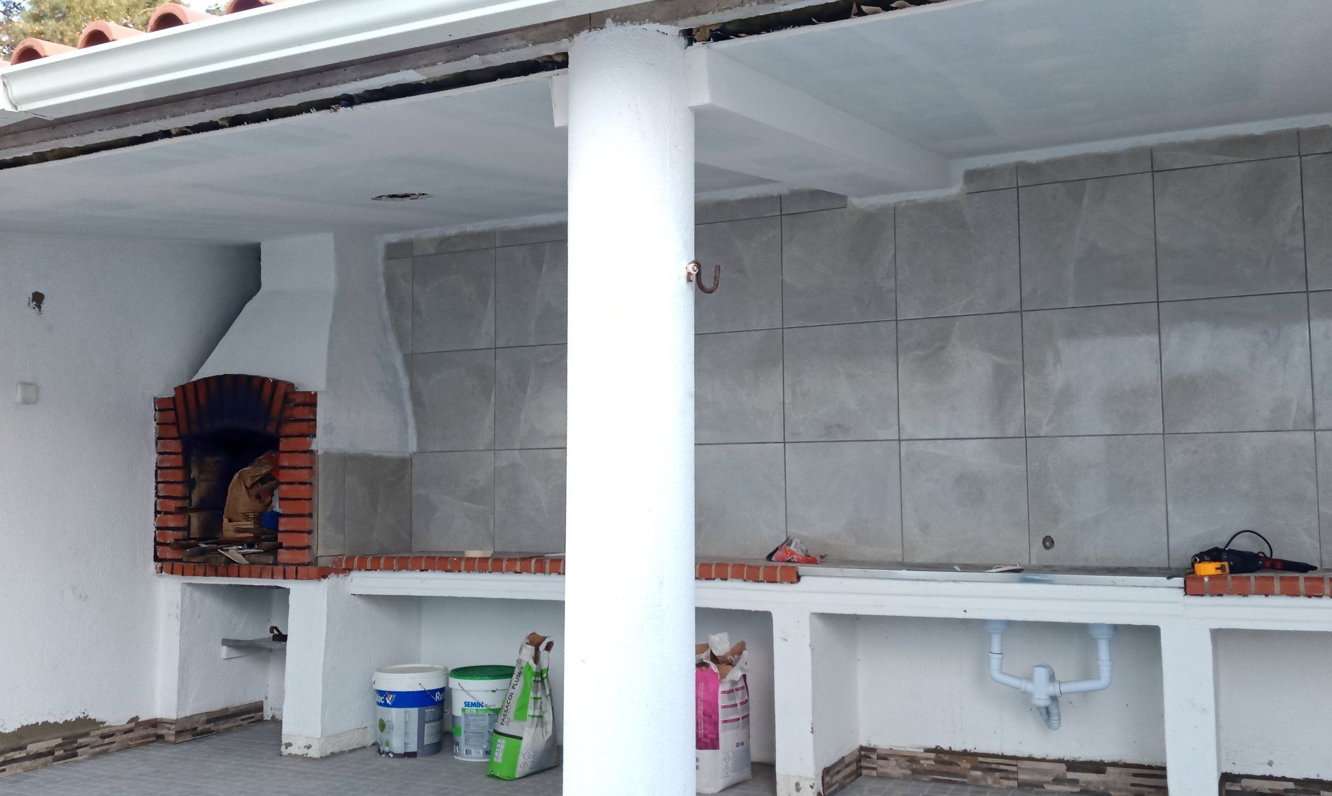 Cozinha externa com churrasqueira de tijolos, bancadas de concreto e parede revestida com azulejos.