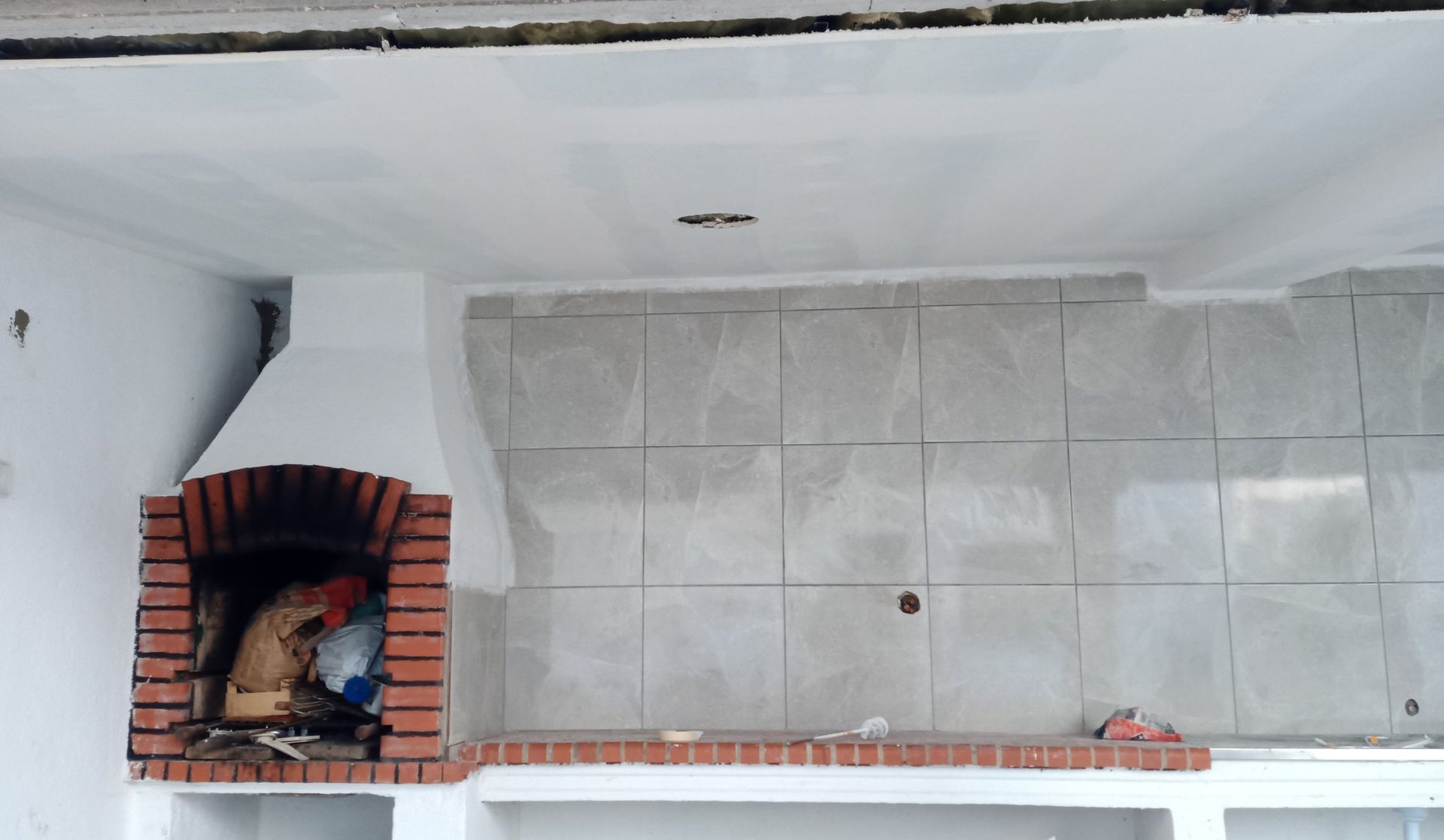 Lareira de tijolos e parede revestida com azulejos em uma cozinha externa de paredes brancas, com bancada e teto brancos.