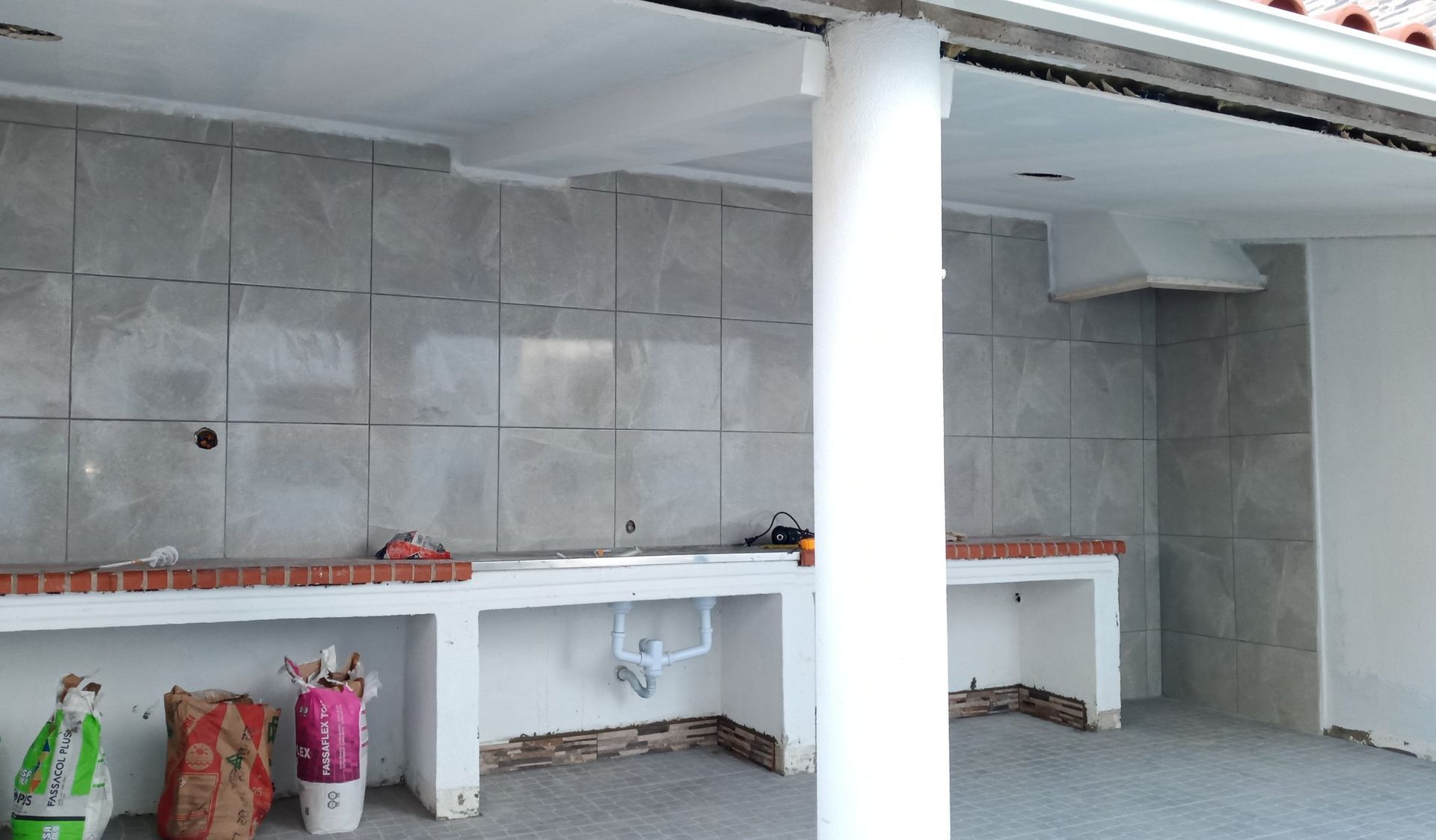 Cozinha externa em construção com paredes revestidas de azulejos cinza e bancadas de concreto.