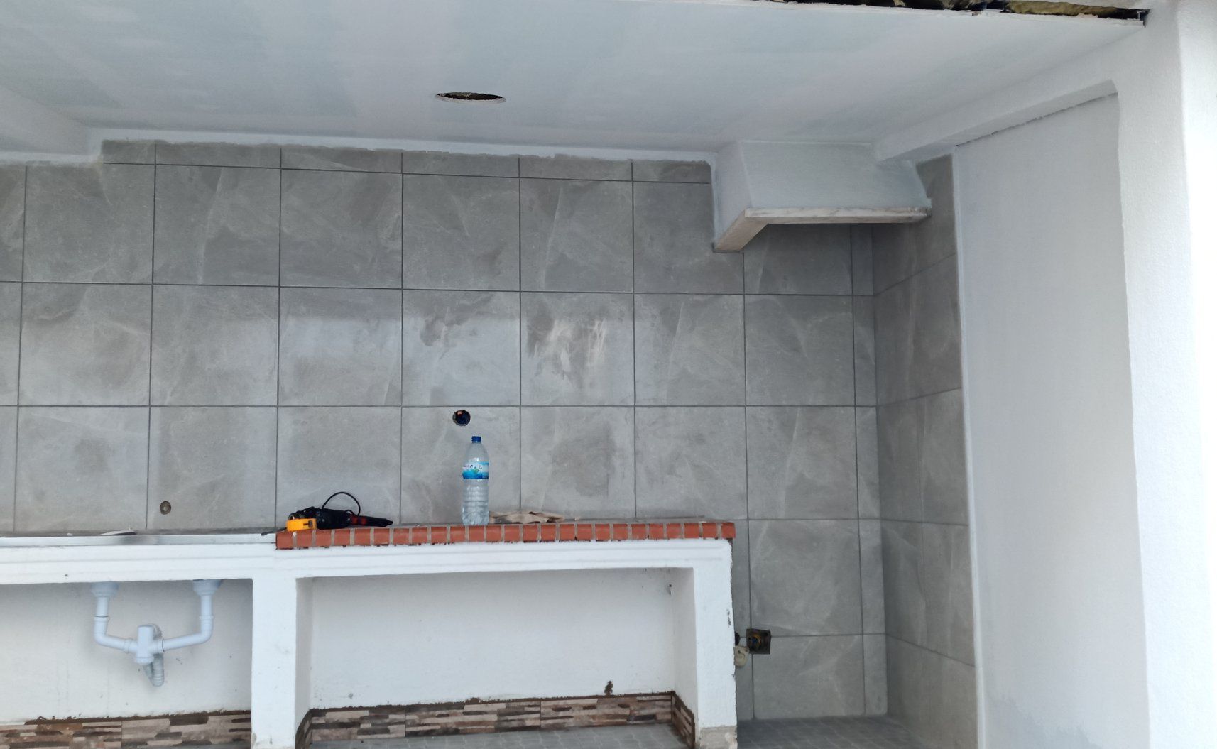 Cozinha em construção com parede revestida de azulejos cinza, bancada branca e encanamento aparente.