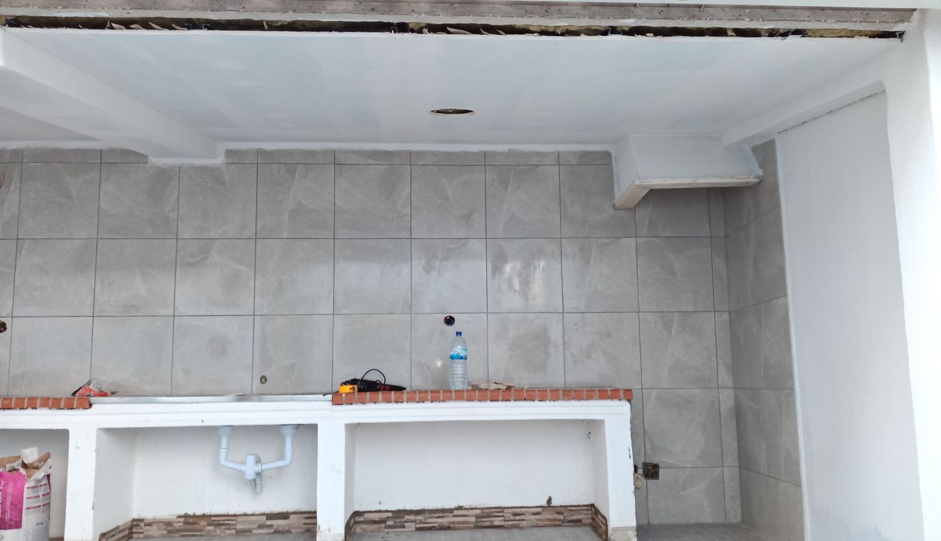Cozinha em construção; backsplash de azulejos cinza, bancadas brancas, teto inacabado.