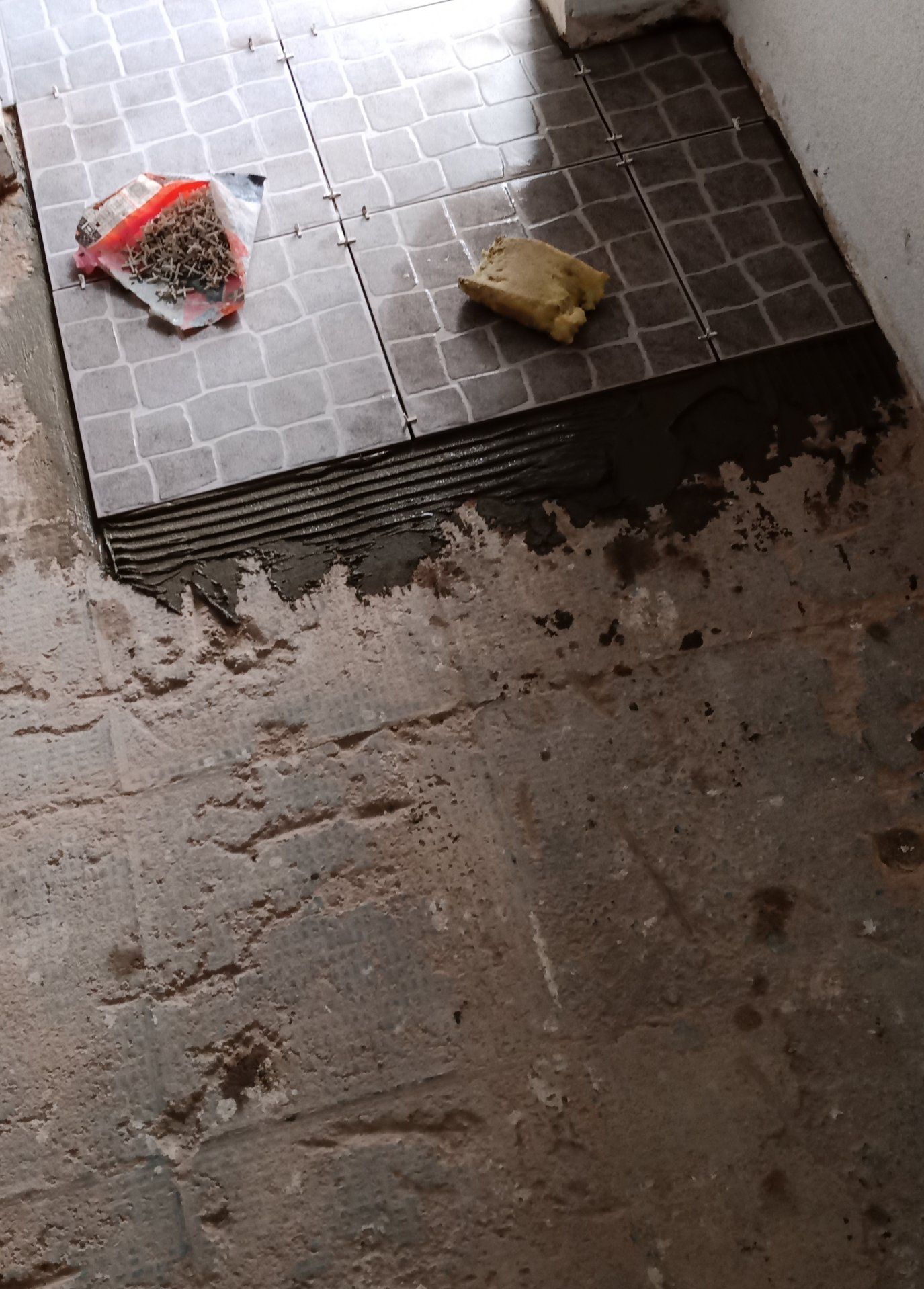 Instalação de novos ladrilhos no piso, parcialmente concluída. Ladrilhos estampados em cinza escuro sobre cimento.