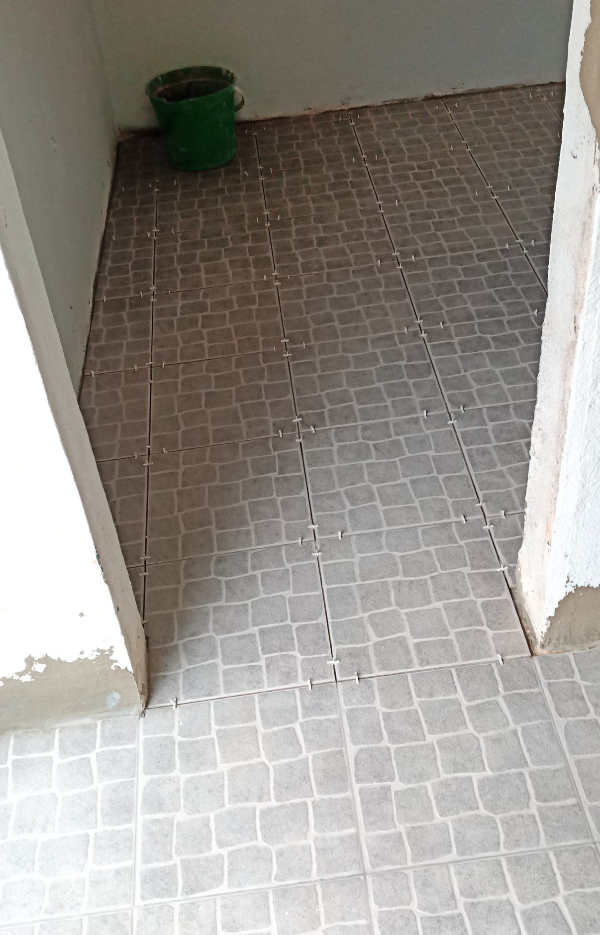 Piso de azulejos na entrada, com um balde verde no canto. As paredes são brancas e o piso é cinza.