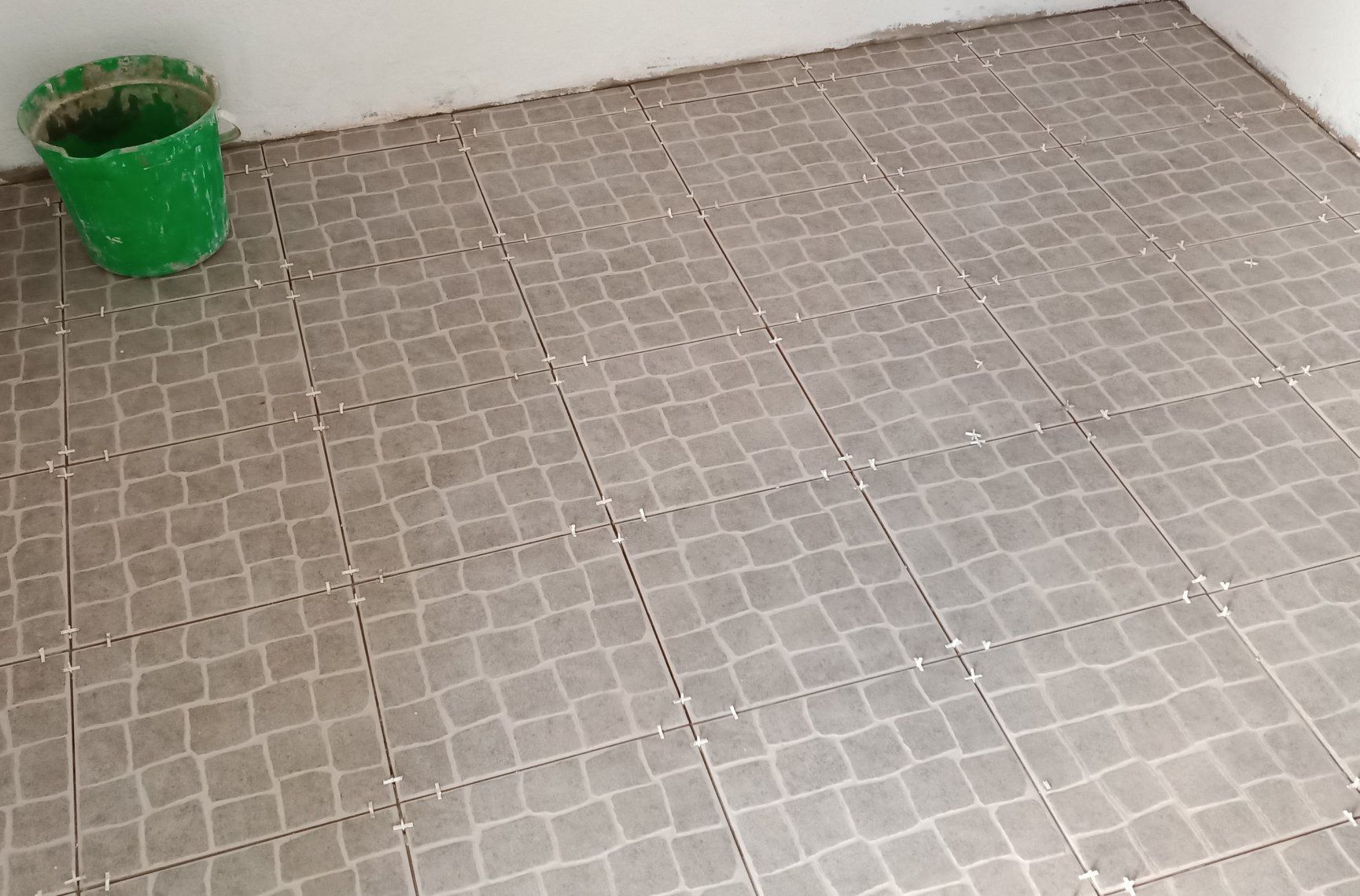Piso de ladrilho cinza com padrão quadriculado e um balde verde em um canto.