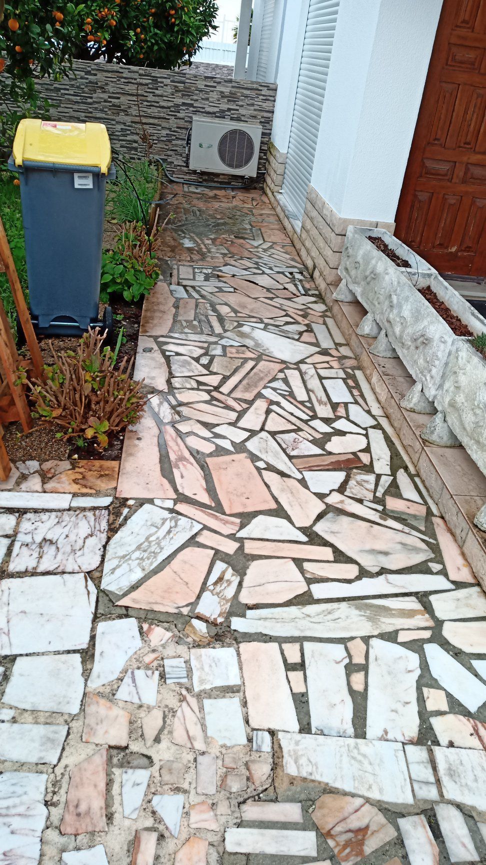 Caminho de pedra ao lado de um prédio branco e uma lixeira com tampa amarela.