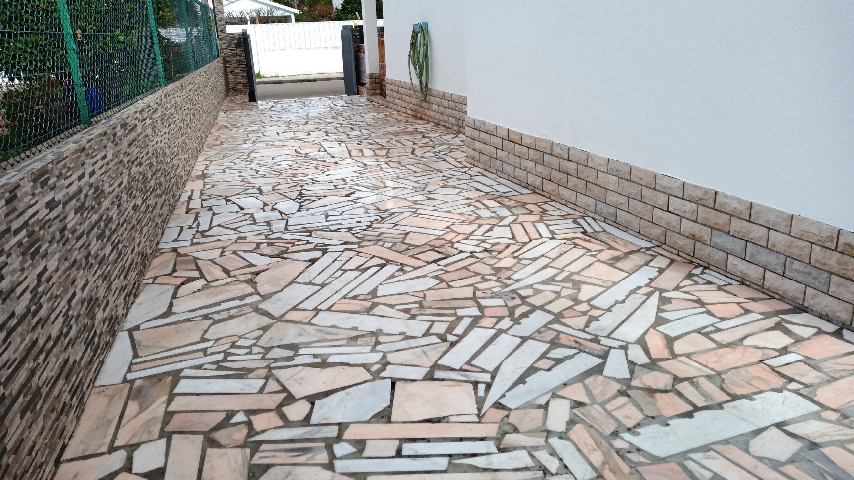 Entrada de automóveis pavimentada com pedras irregulares de cor clara, delimitada por pedra e um muro branco, que leva a um portão.