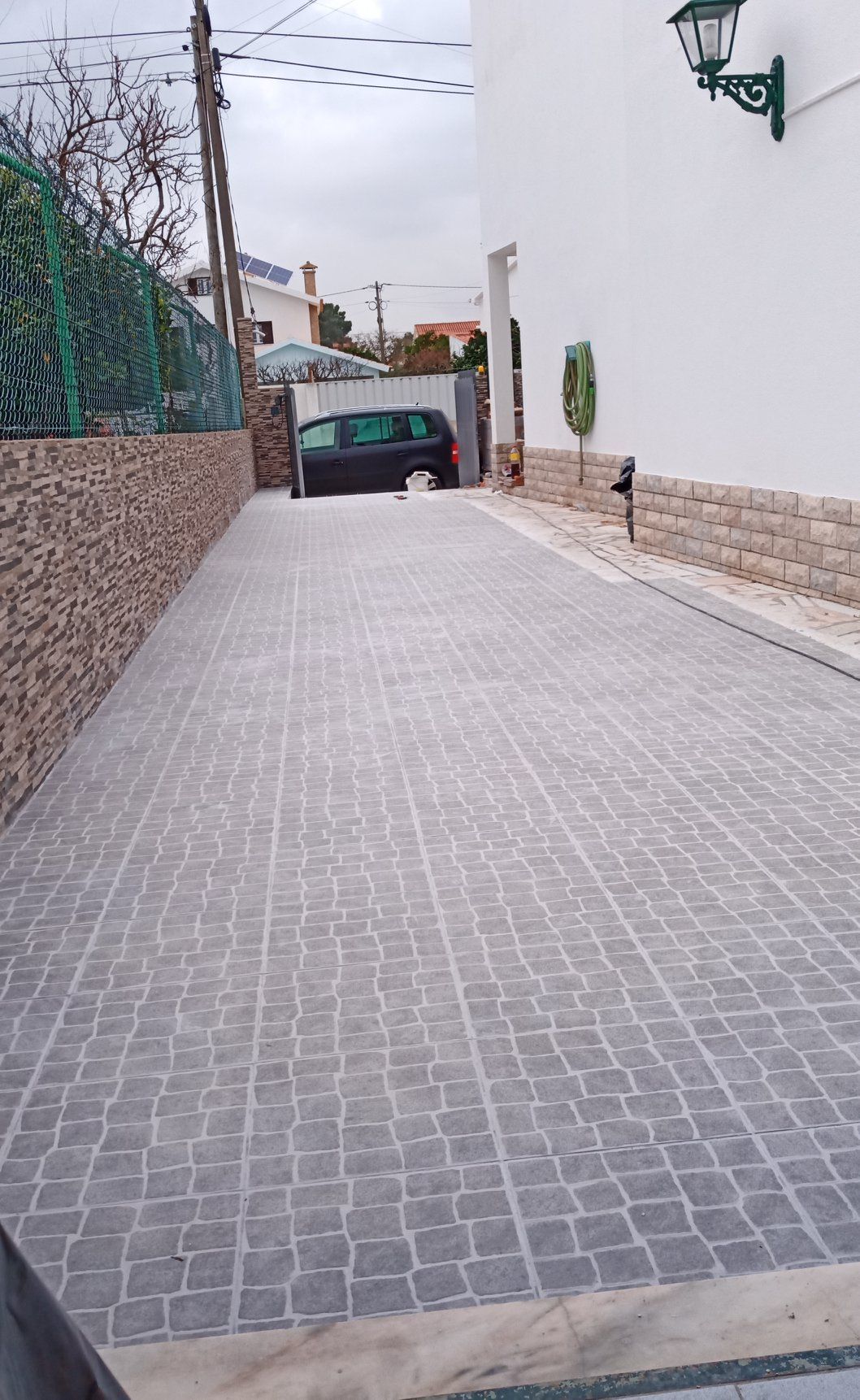 Entrada de automóveis pavimentada com paralelepípedos que leva a uma garagem com um carro dentro; prédio branco à direita, cerca viva verde à esquerda.