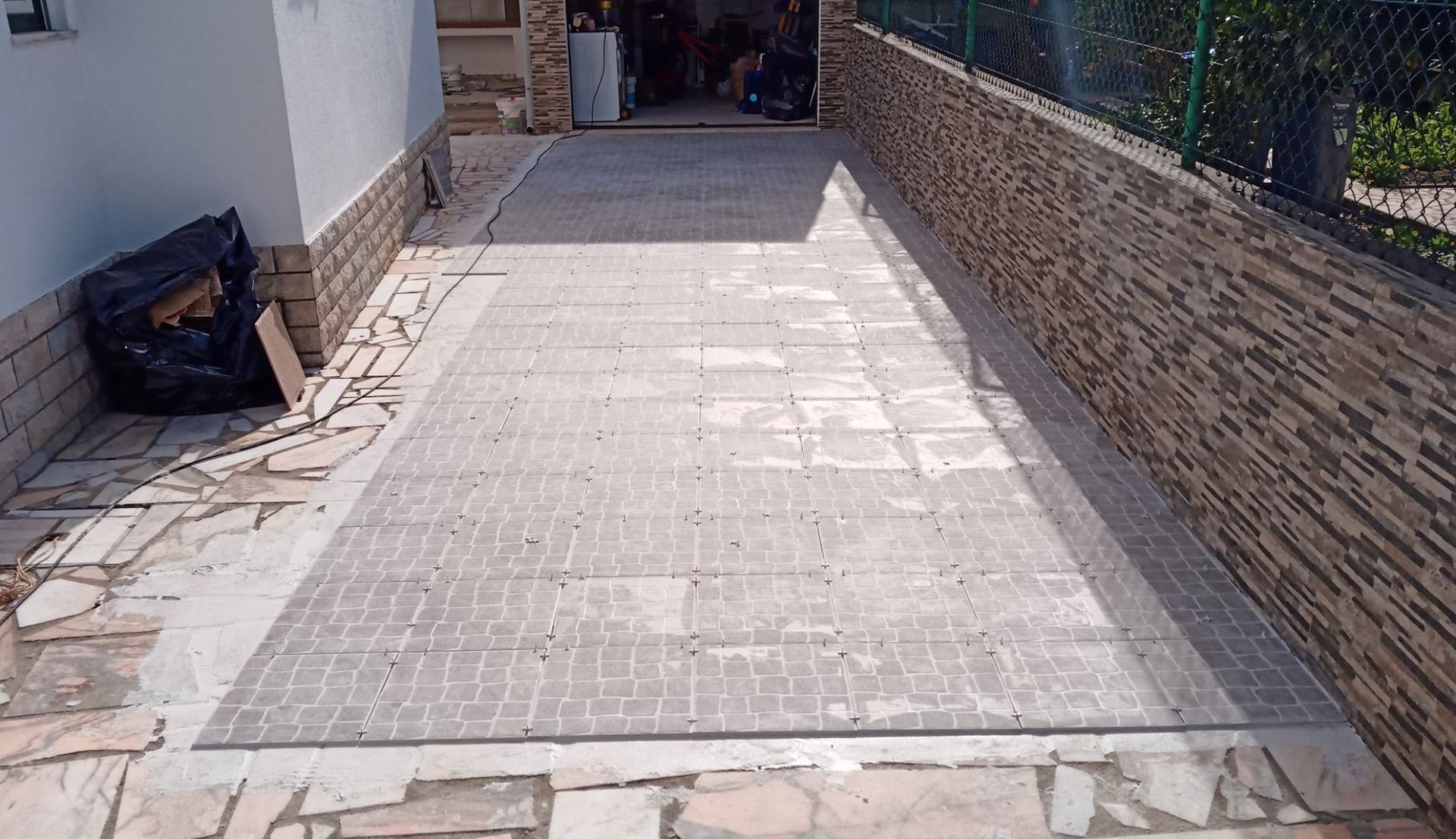 Entrada de automóveis em concreto com padrão semelhante a tijolos, ladeada por um muro de pedra e um prédio com um saco de lixo preto.