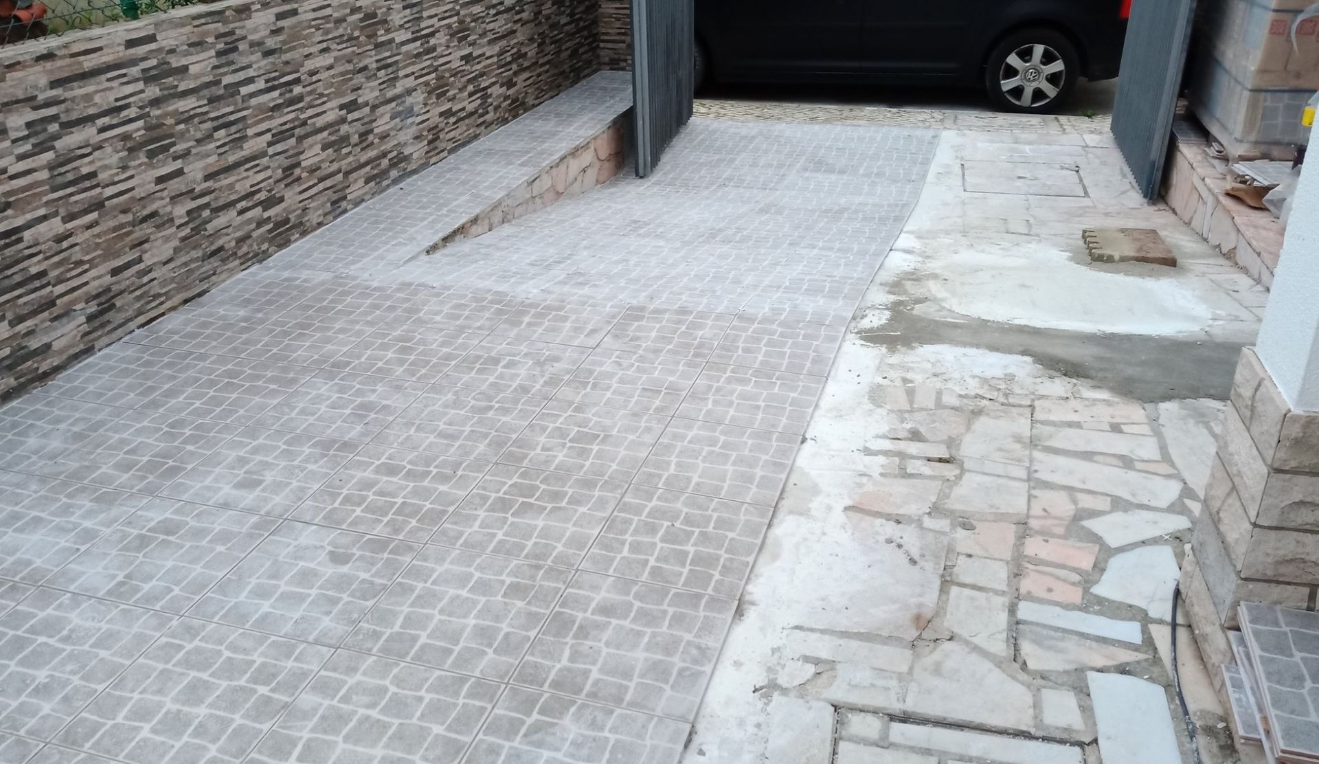 Entrada de automóveis em concreto com muro de pedra, que leva à garagem. Um carro cinza está estacionado dentro dela.