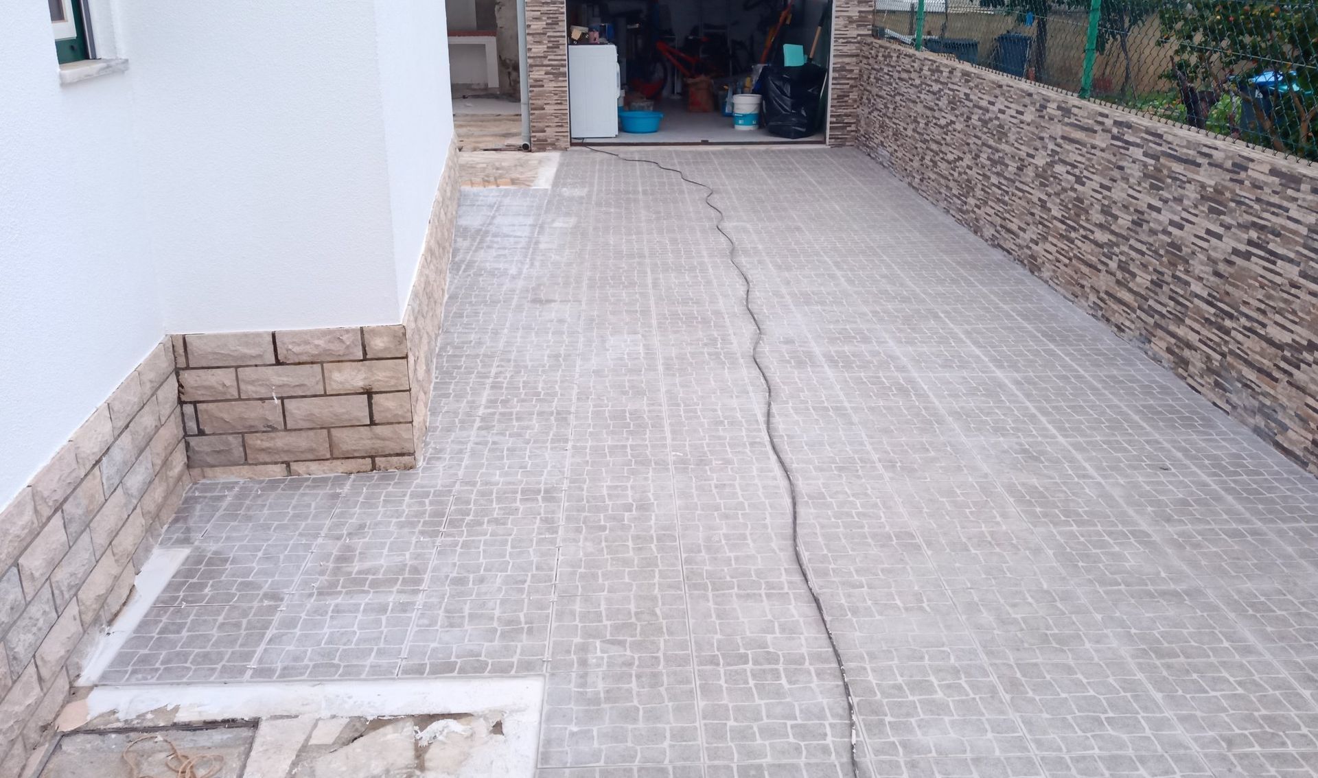 Entrada de automóveis em concreto com uma grande rachadura, delimitada por muros de tijolo e pedra, que dá acesso a uma garagem aberta.