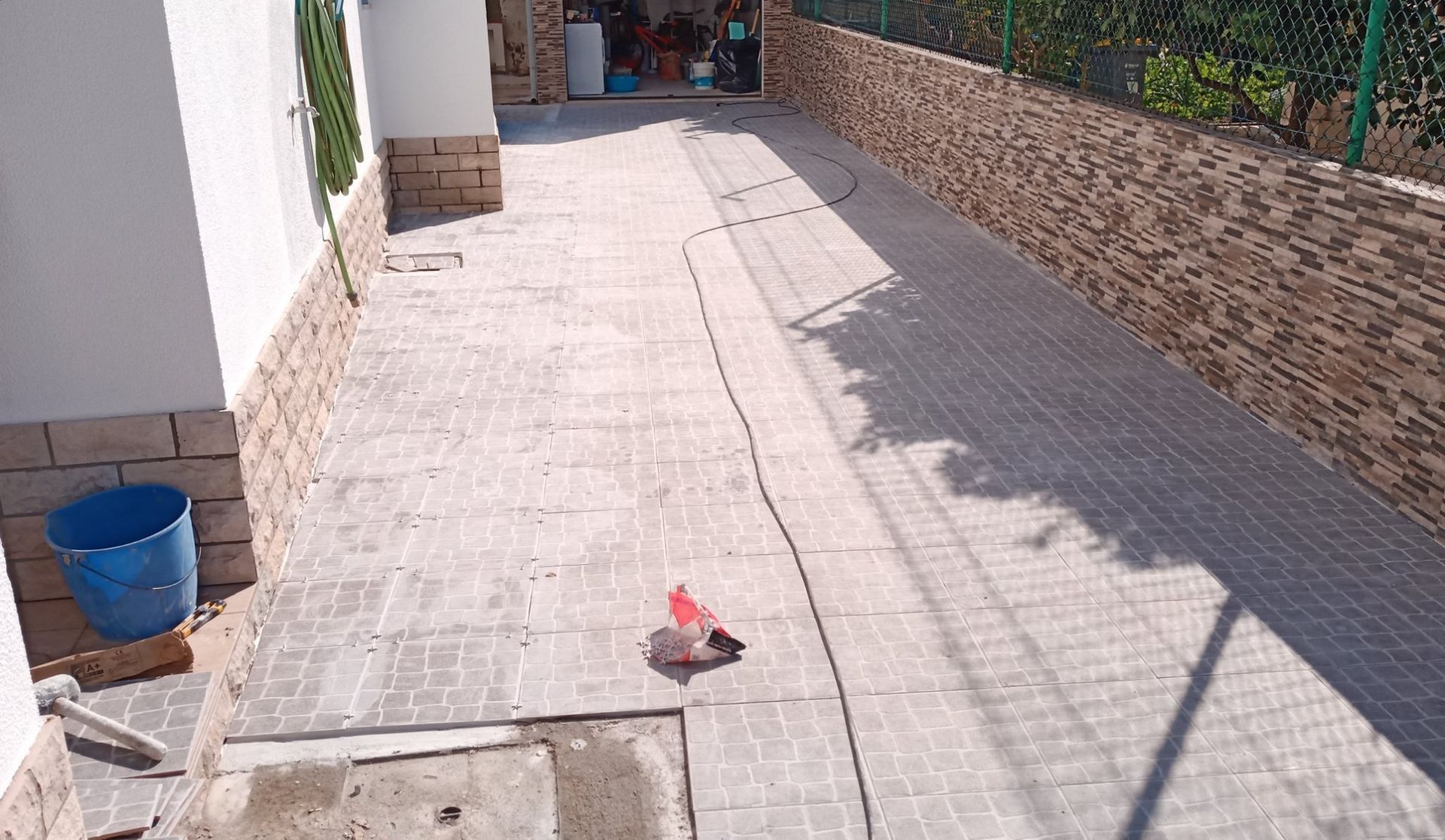 Uma estreita passarela pavimentada com um muro de pedra e um edifício de cada lado. Um balde azul e detritos são visíveis.