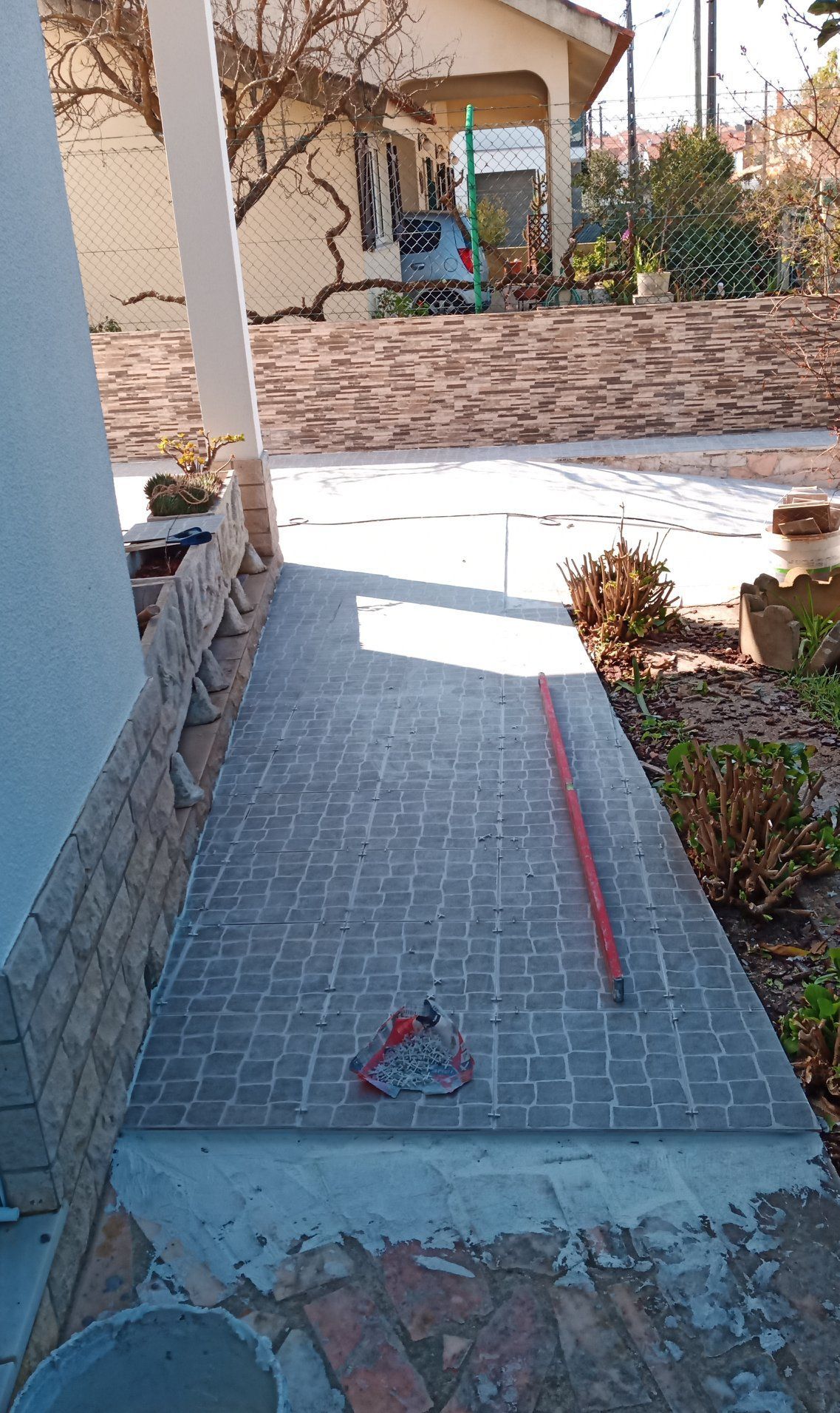 Caminho pavimentado com tijolos cinza-escuros, que leva a uma casa; ferramentas estão sobre o caminho.