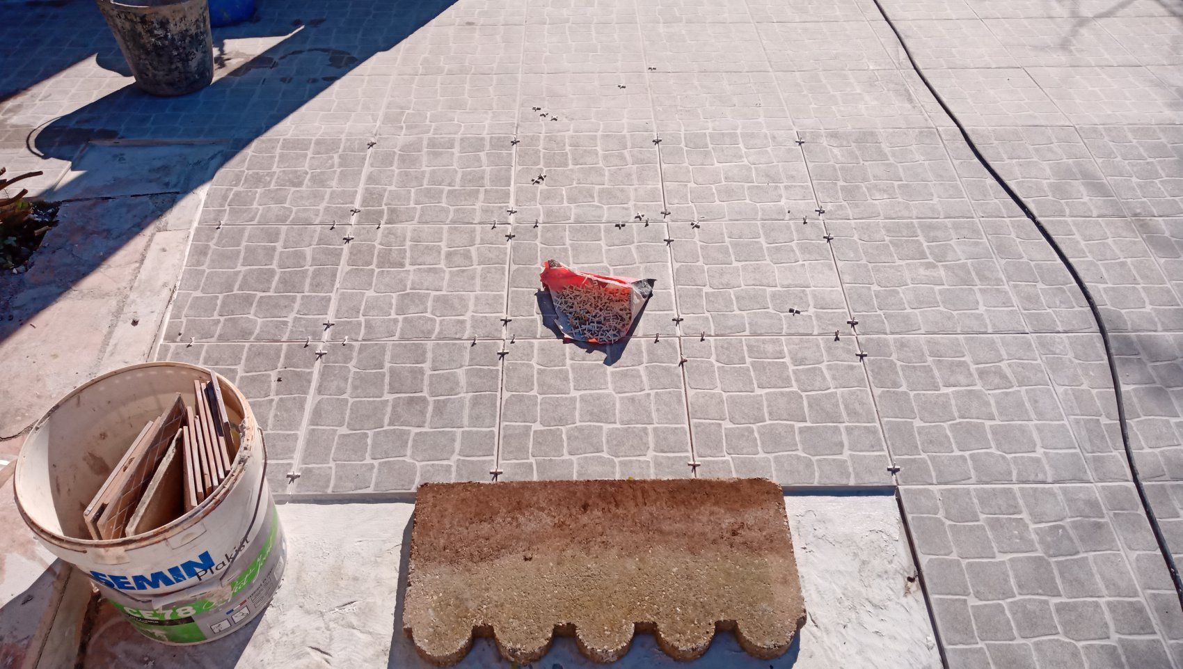 Calçamento de paralelepípedos em andamento, apresentando um balde de ladrilhos, um objeto vermelho e ferramentas sobre pedras cinzentas.