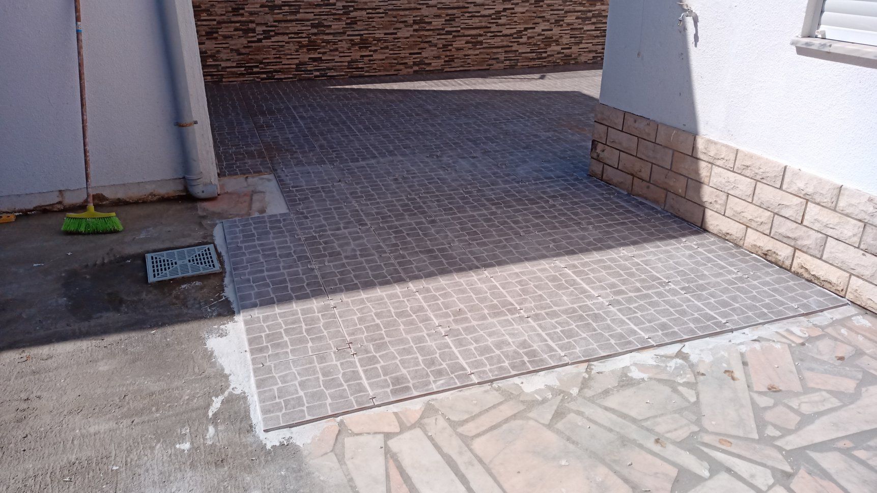 Caminho de tijolos que liga uma área pavimentada à entrada de um edifício. Uma vassoura está encostada na parede.