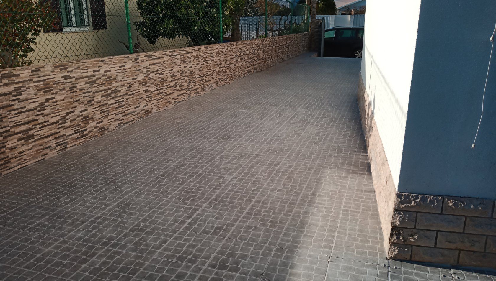 Longa entrada pavimentada, delimitada por um muro de tijolos texturizados e um prédio branco; luz solar.