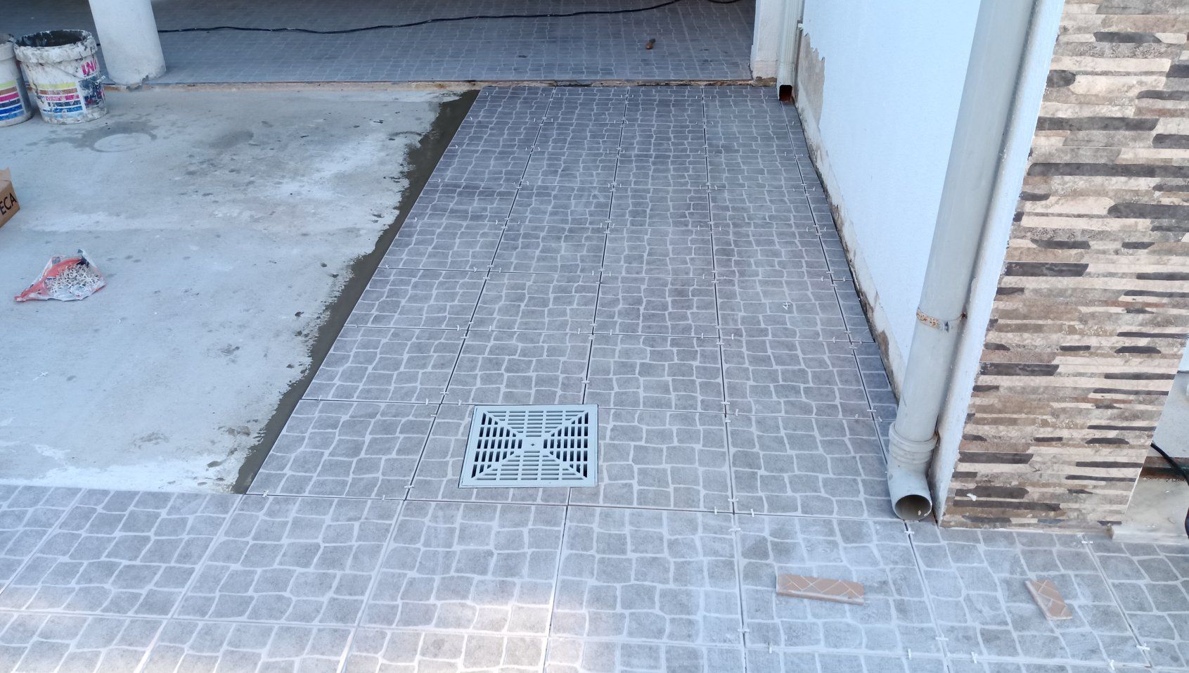 Entrada de automóveis em paralelepípedos que leva a uma garagem, com um ralo e uma faixa de cimento.