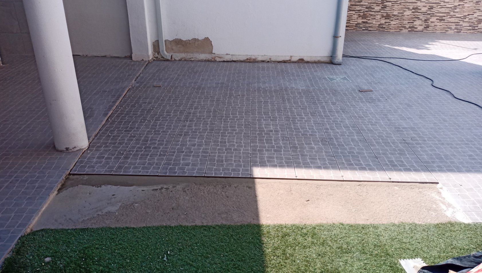 Pátio com piso de azulejo cinza, grama artificial e um pilar branco.