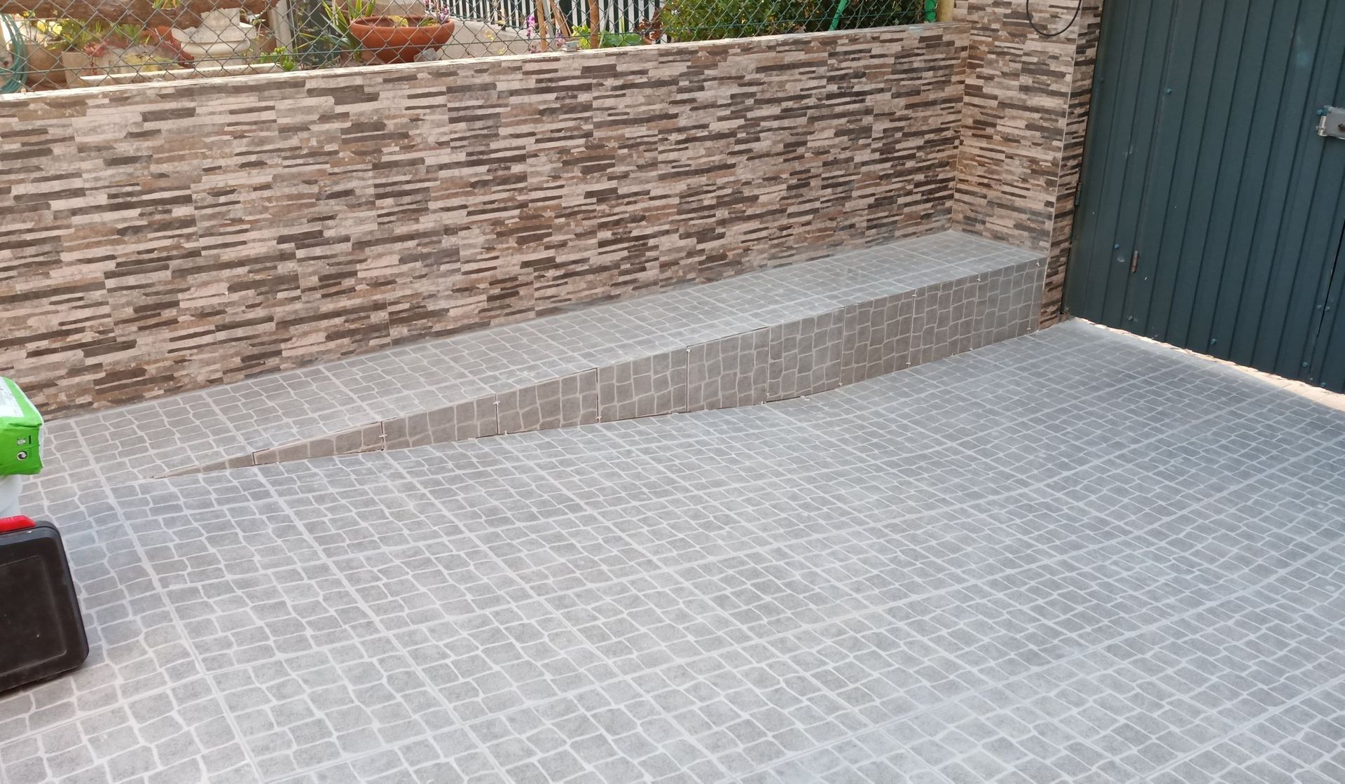 Uma rampa revestida com ladrilhos cinzentos conduz a uma porta verde, construída num edifício com tijolo castanho texturizado.