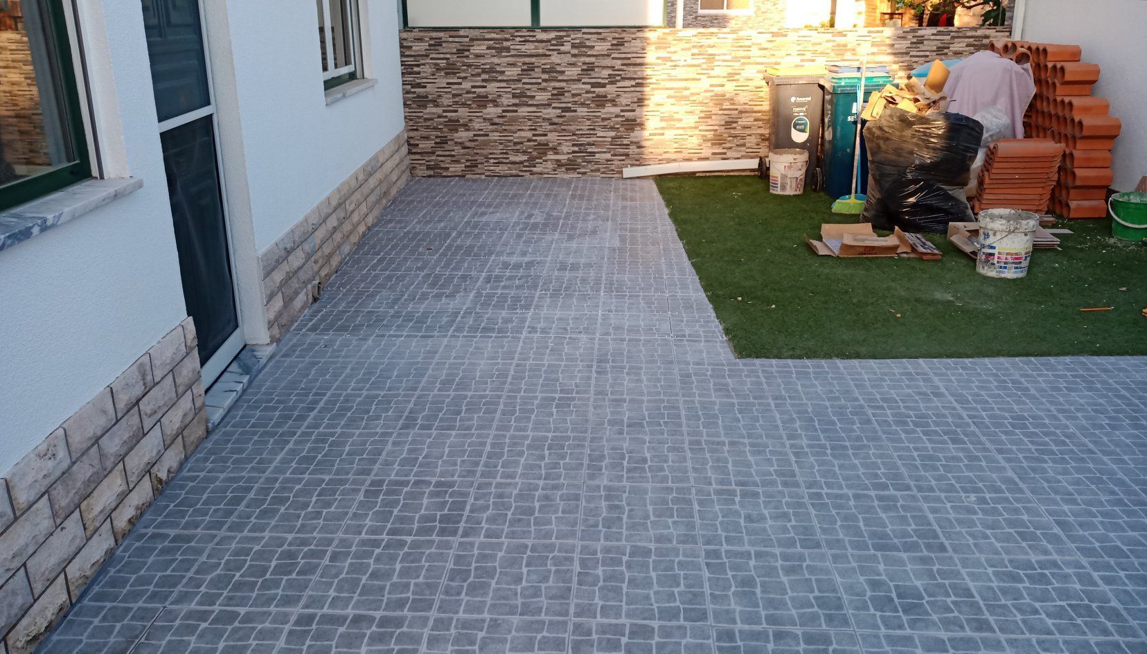 Pátio pavimentado com tijolos cinza, com transição para grama artificial. Uma pessoa está trabalhando ao fundo.