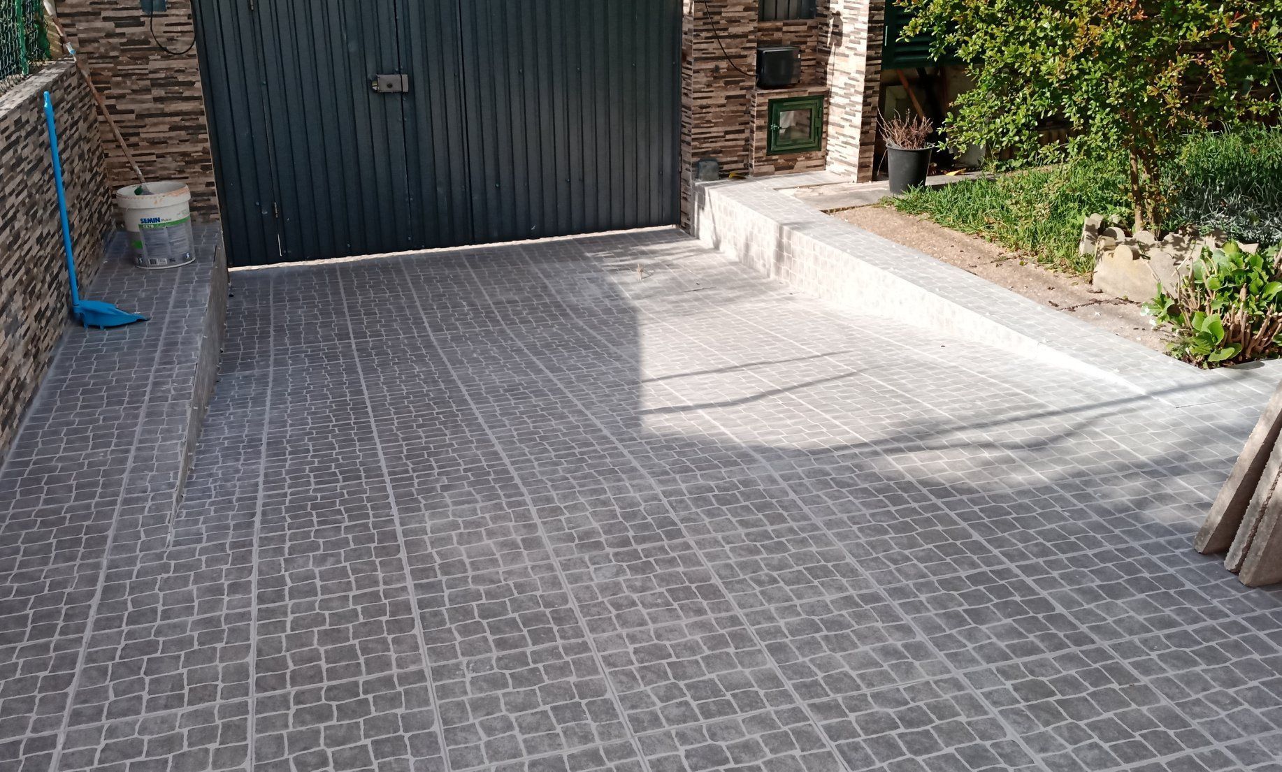 Entrada de automóveis em tijolo escuro que leva a um portão preto fechado; muros de pedra e vegetação cercam o local.