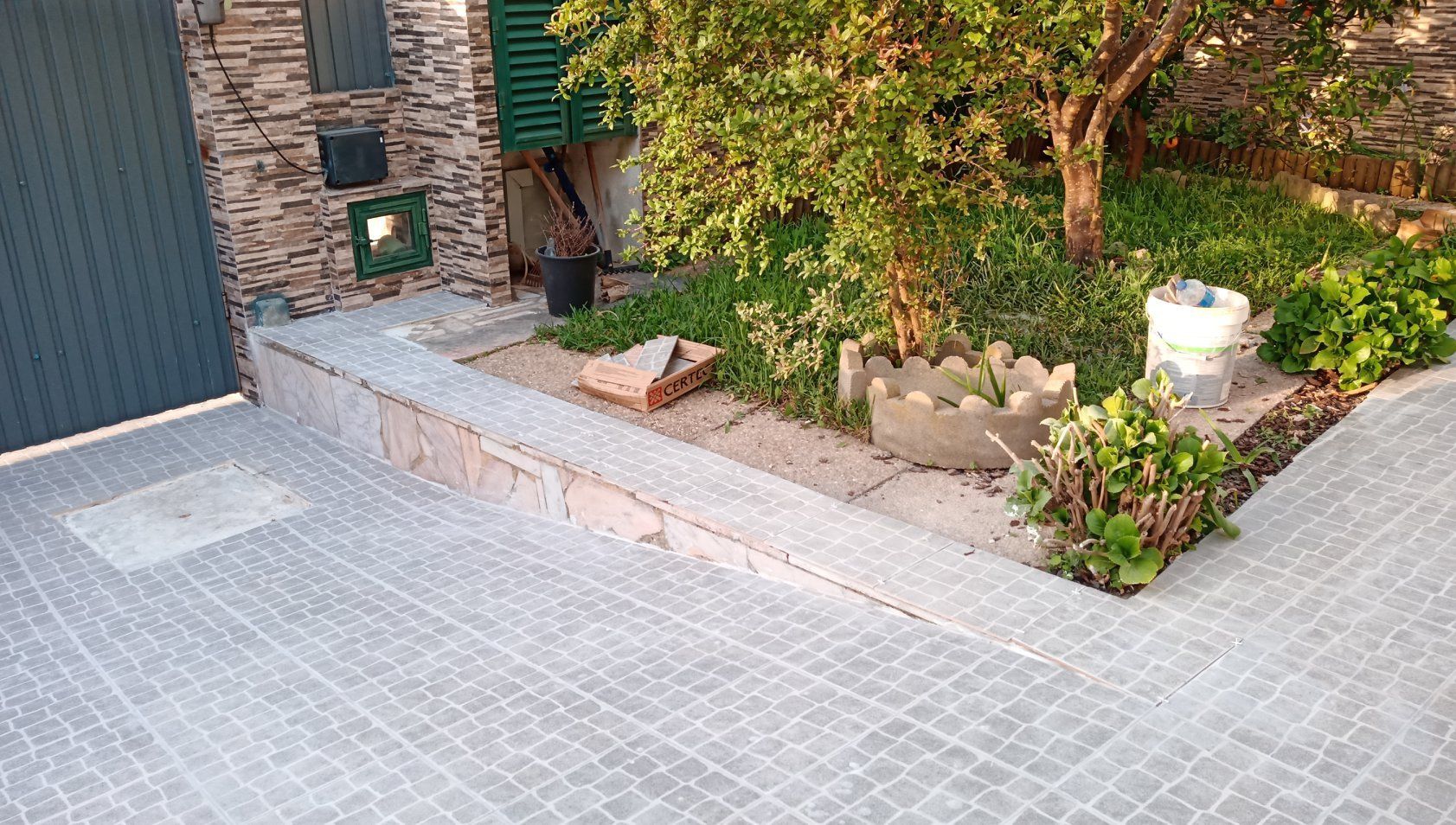 Entrada de automóveis pavimentada em cinza ao lado de um pequeno jardim com arbustos verdes e uma árvore.