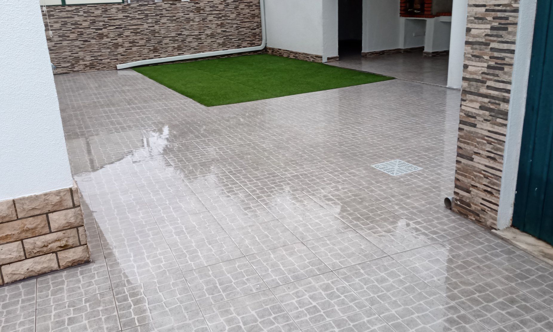 Pátio com ladrilhos estampados e molhados. Pequeno trecho de grama artificial verde. Paredes com detalhes em tijolo aparente.