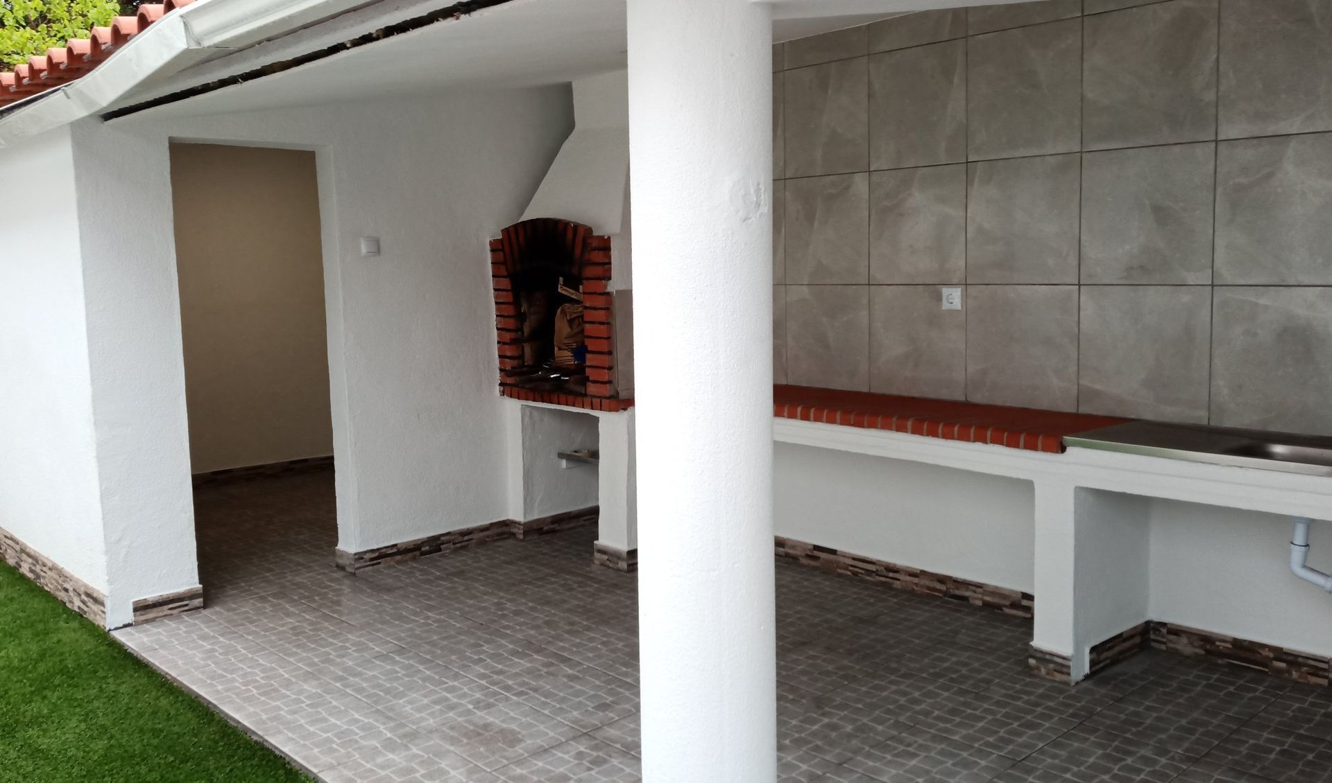 Cozinha externa coberta com churrasqueira de tijolos, balcão e pilar branco.