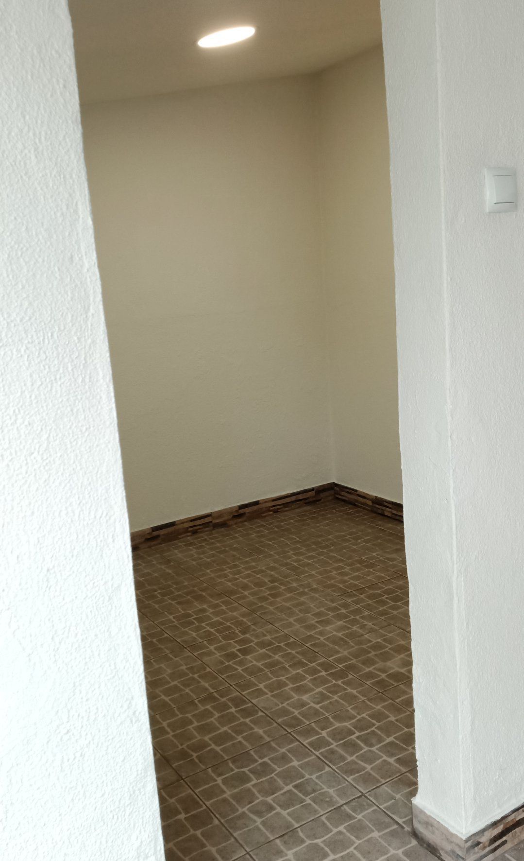 Quarto vazio com paredes branco-sujo, piso de azulejo e uma única lâmpada no teto, visto através de uma abertura.